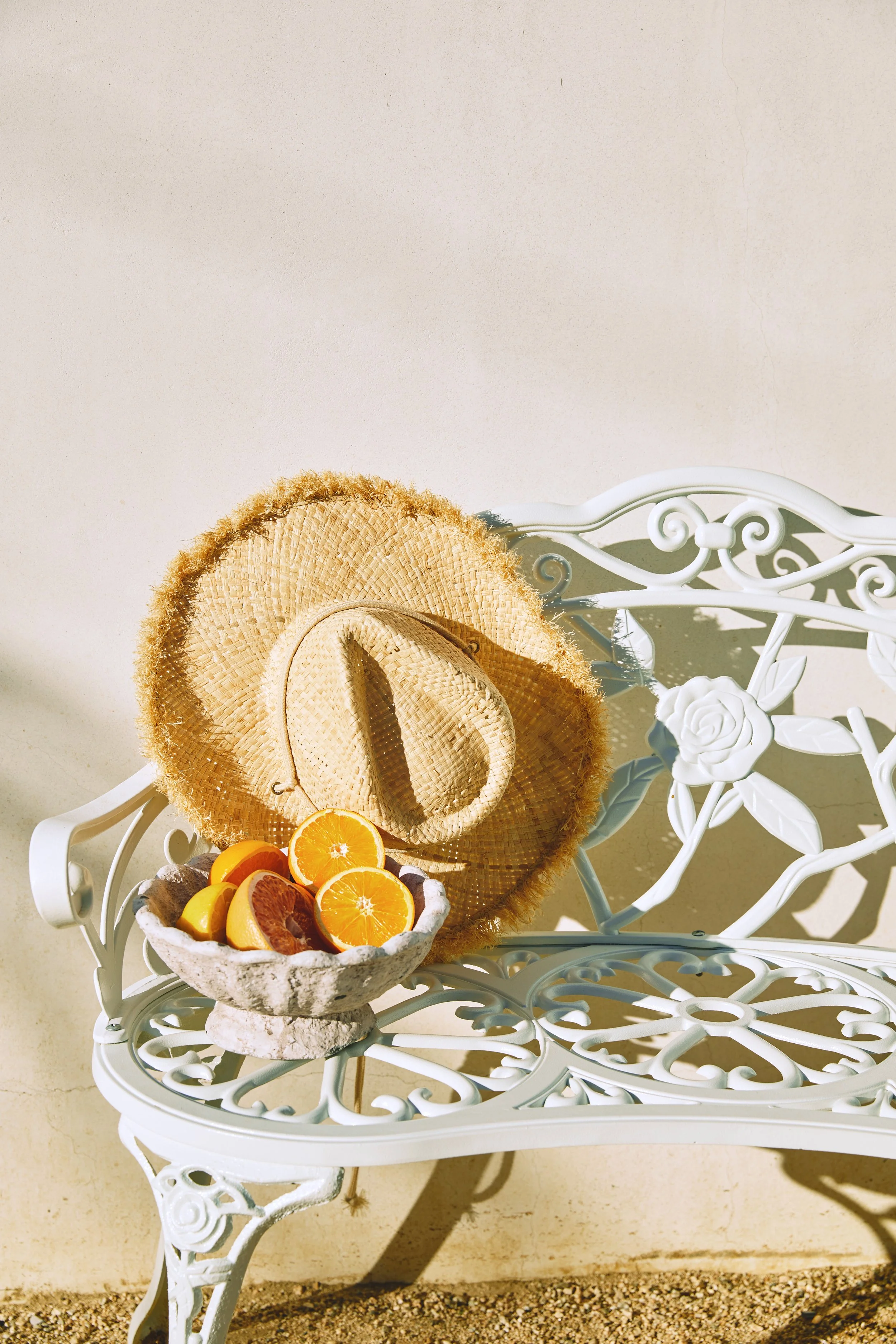 A white decorative bench with a floral design, a straw sun hat, and a bowl of sliced oranges, lemons, and a blood orange, set against a light-colored wall.