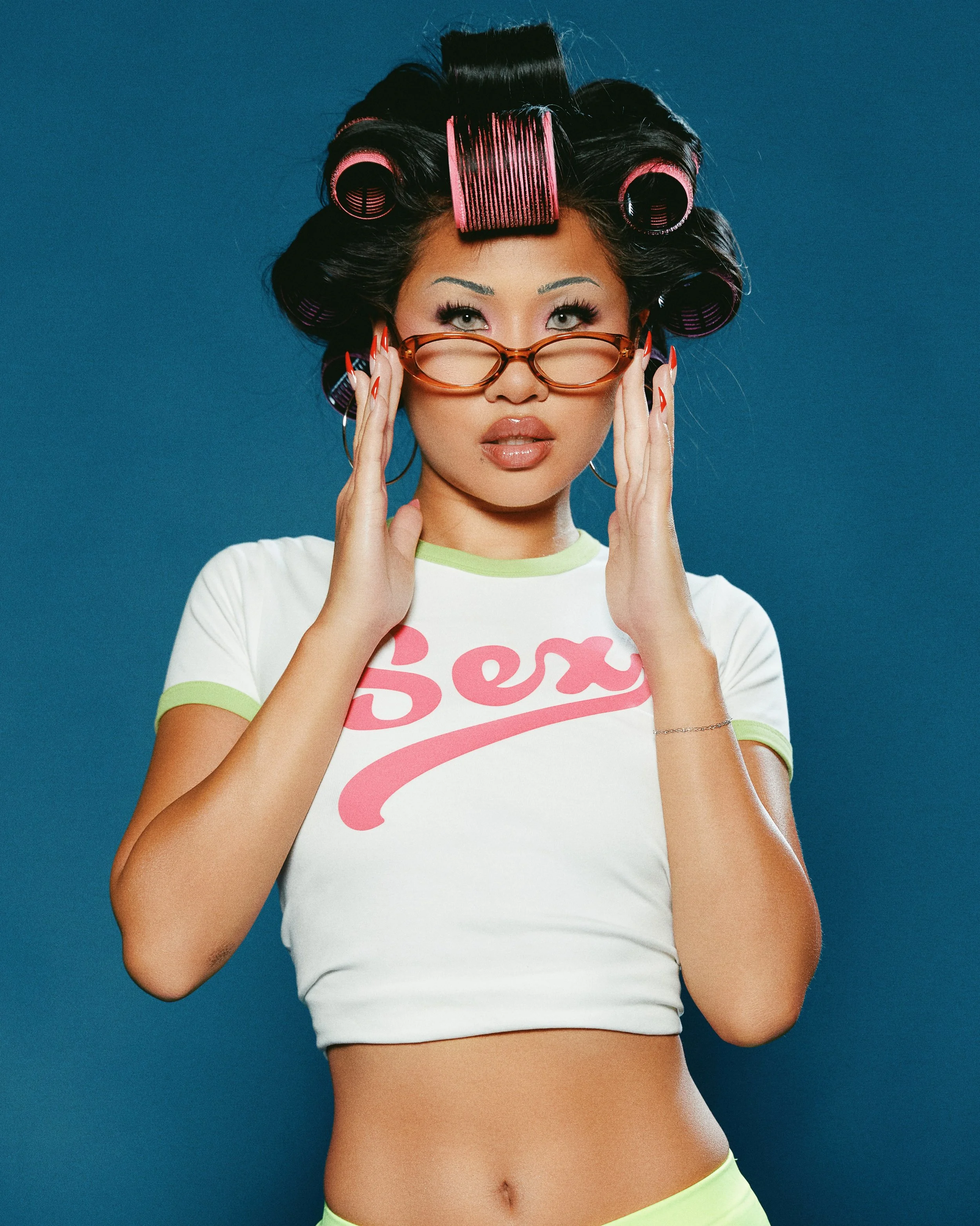 A woman with hair rollers, wearing glasses, and a white t-shirt with pink letters spelling 'Sex' against a blue background.