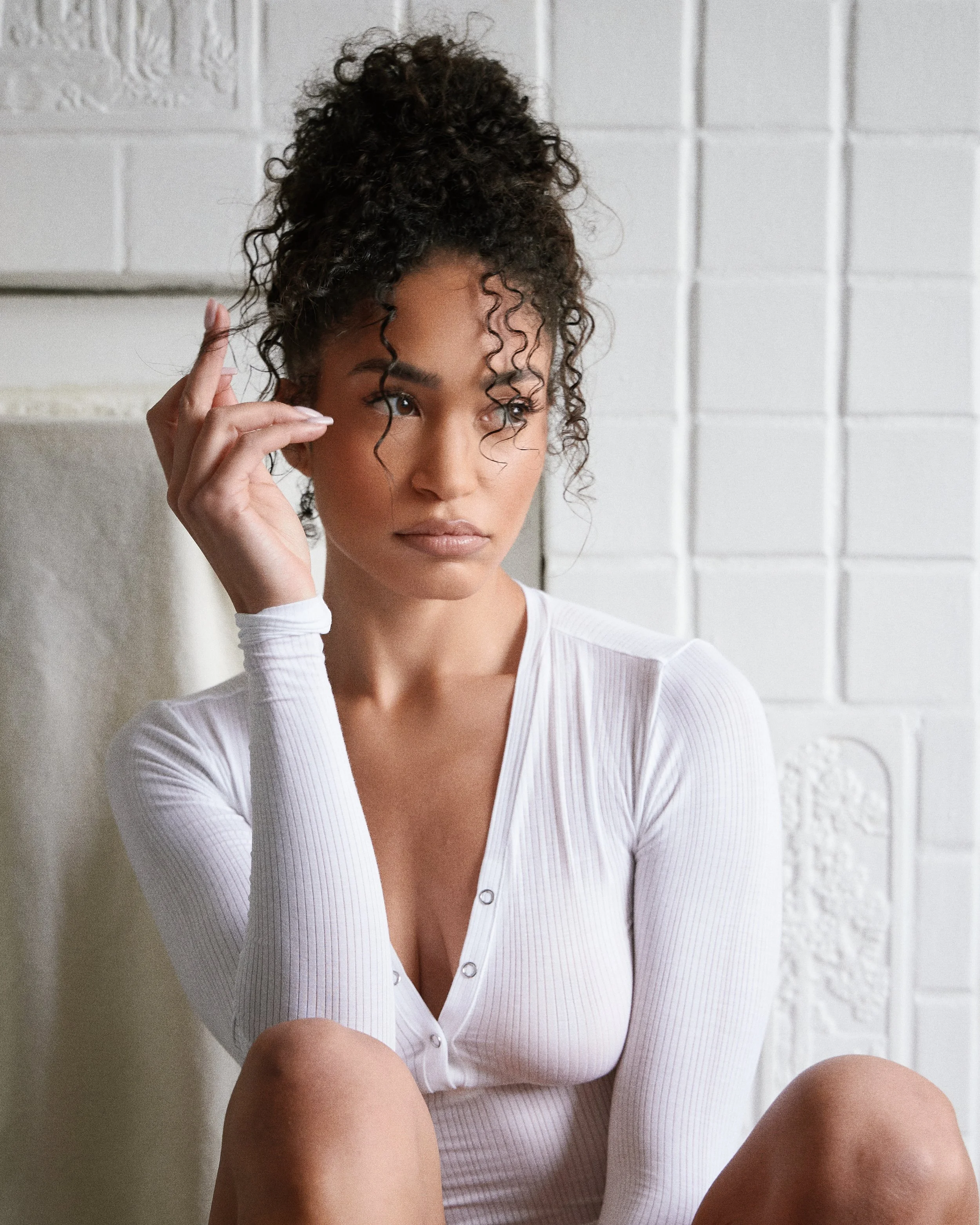 A woman with curly hair tied up, wearing a white ribbed long-sleeve top with a deep neckline, sitting against a white brick wall.