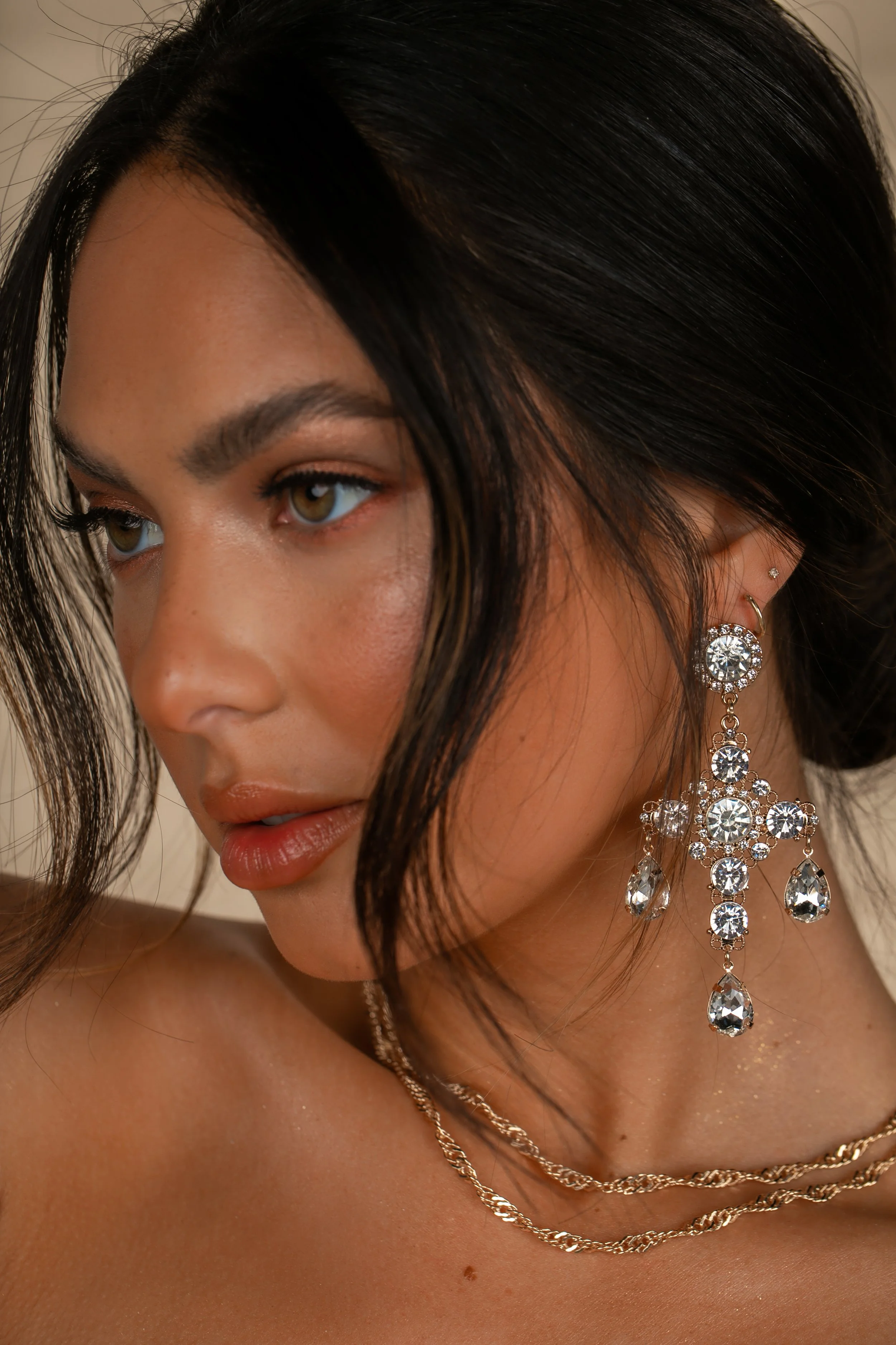 Close-up of a woman with dark hair and hazel eyes wearing sparkling diamond earrings and layered gold necklaces.