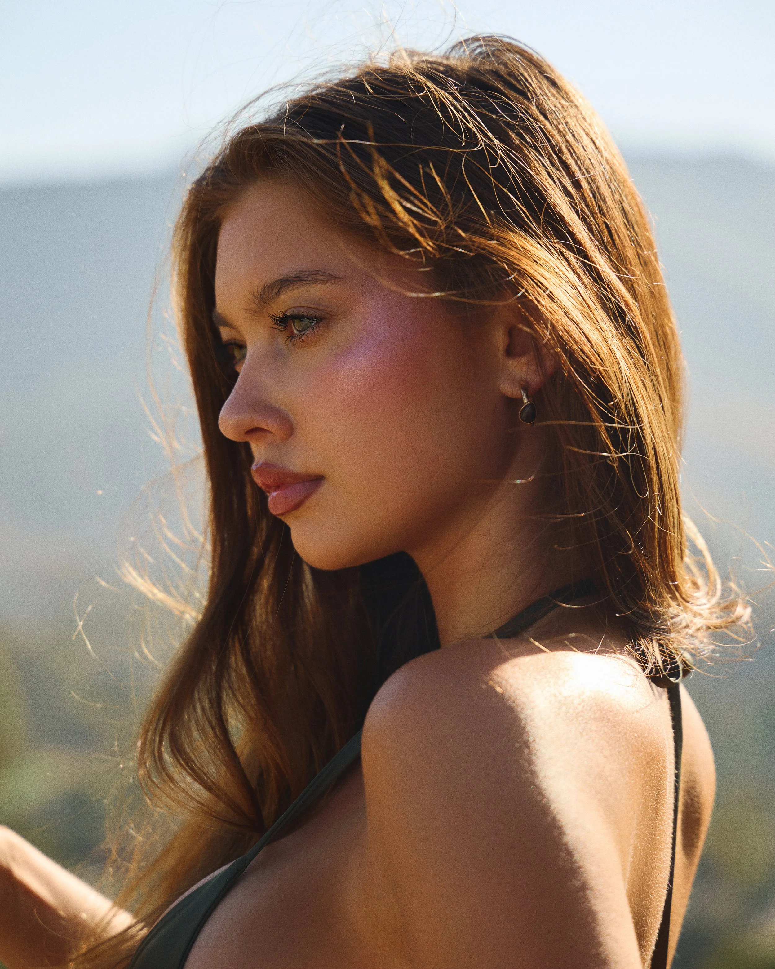 Profile of a young woman with long, auburn hair, wearing earrings and a tank top, looking away outdoors during daylight.