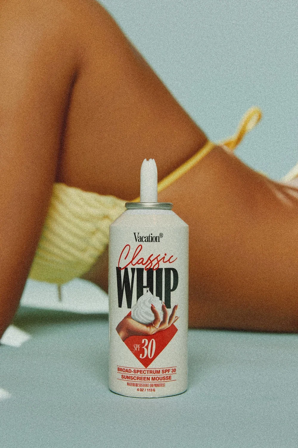A bottle of Vacation Classic Whip sunscreen mousse SPF 30 with a white and red label, placed on a flat surface with a person's arm and yellow swimsuit in the background.