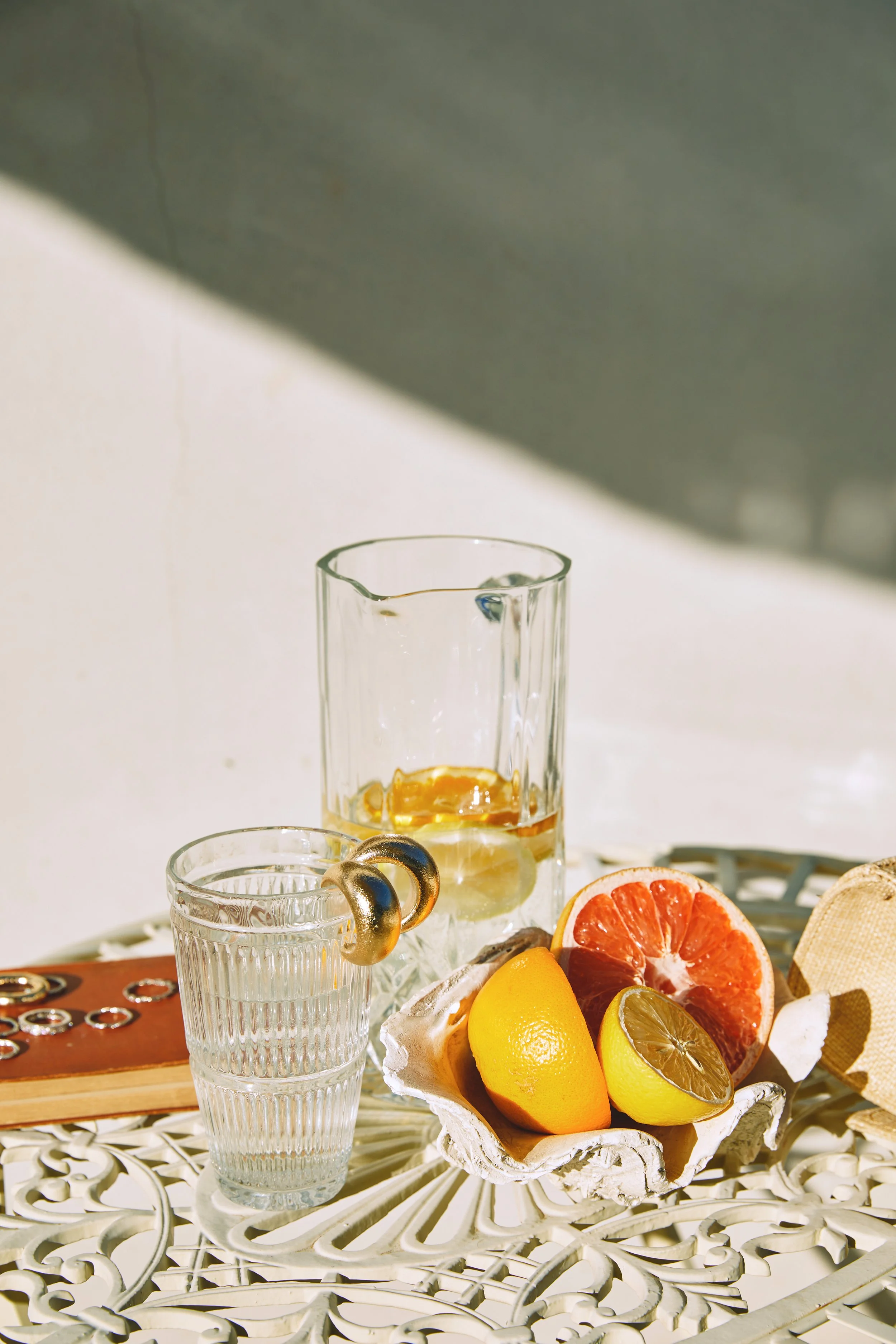 Assorted citrus fruits including lemon, lime, orange, and grapefruit on a decorative white table, with glasses of water and a drink with ice and lemon slices, in natural sunlight.