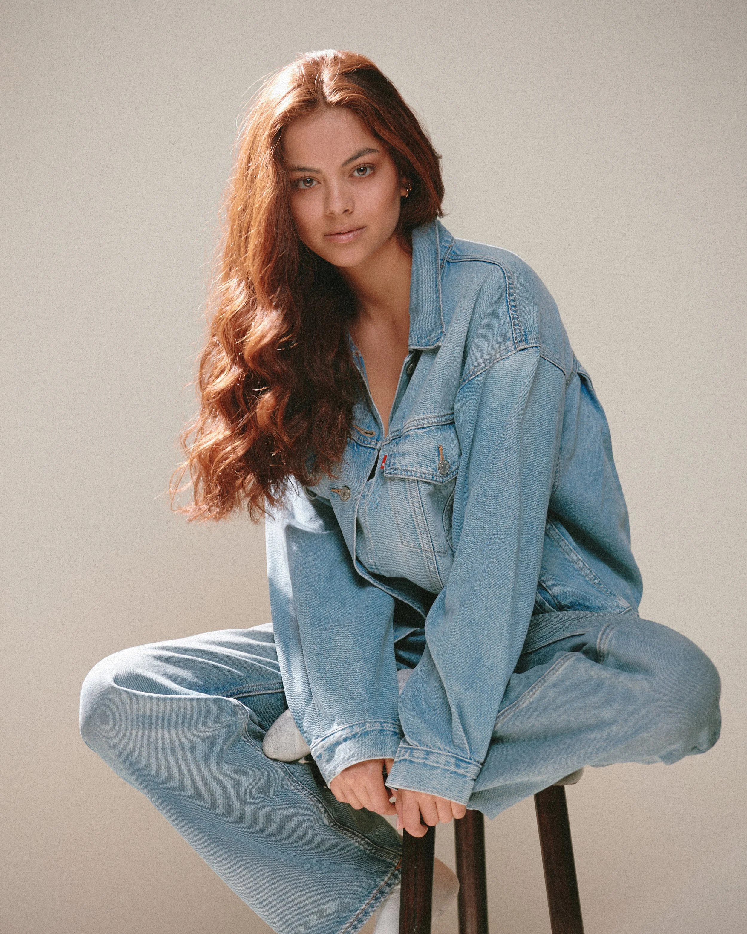 A young woman with long, wavy, reddish-brown hair sitting cross-legged on a stool against a plain, light background. She is wearing a light blue denim jacket and matching jeans, and looking at the camera.