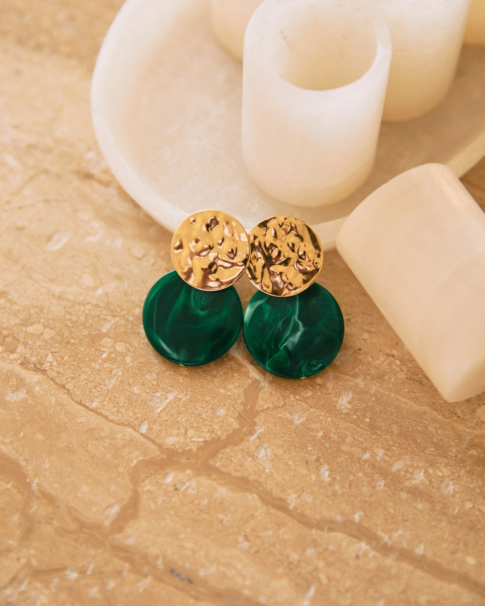 Pair of stylish earrings with gold textured tops and dark green marbled bottoms, placed on a stone surface alongside white candles.