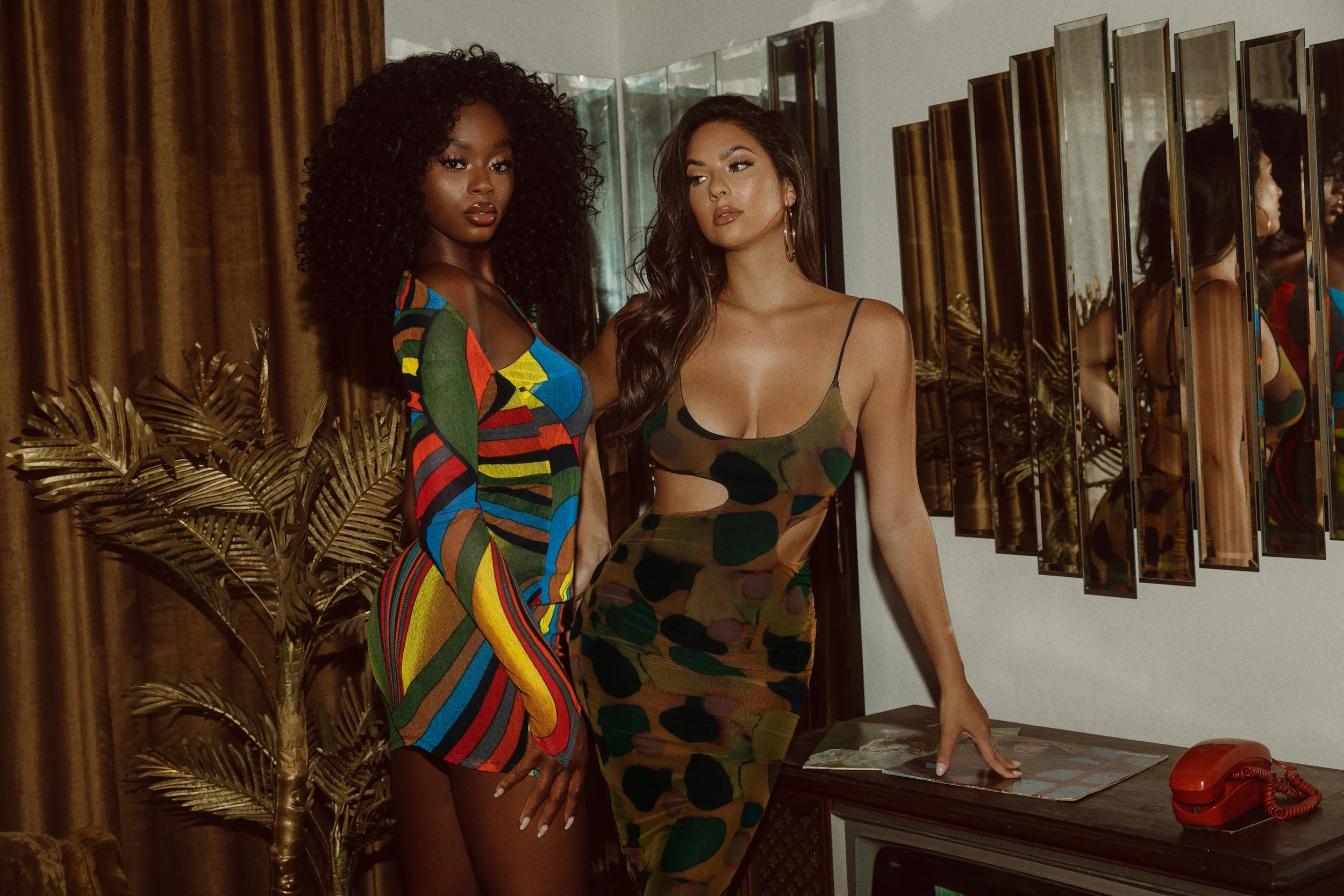 Two women standing in an indoor room with mirrors, a vintage television, a red rotary phone, a decorative gold palm, and gold curtains. One woman has curly black hair and is wearing a colorful, patterned dress. The other woman has long brown hair and