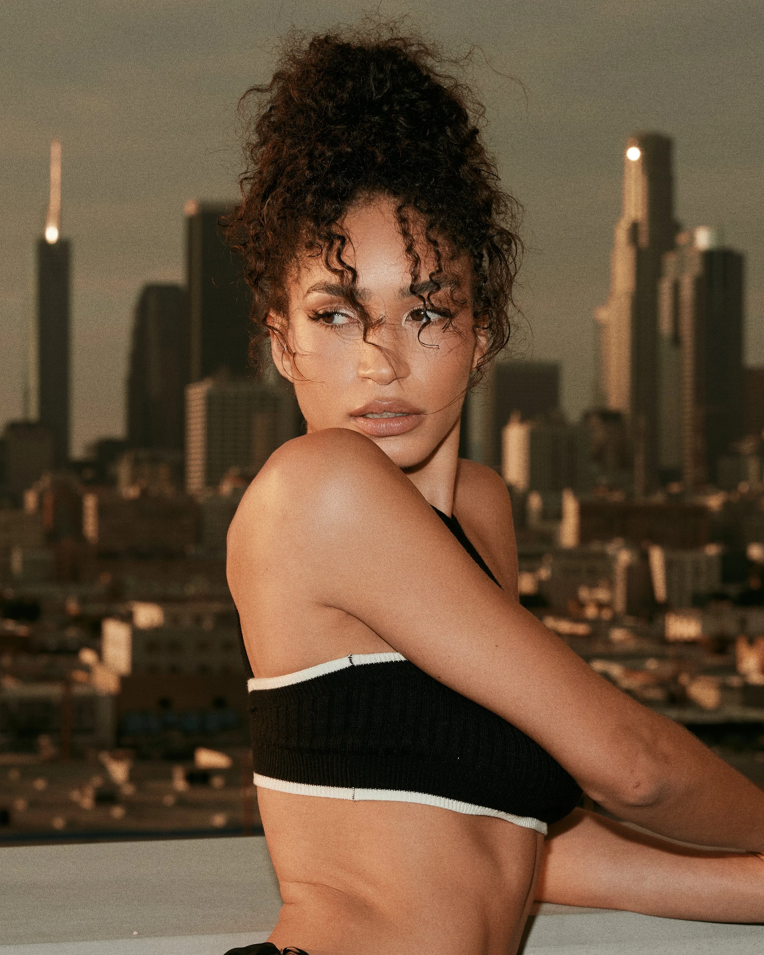 A woman with curly hair looking to the side, dressed in a black and white top, with a city skyline in the background during dusk.