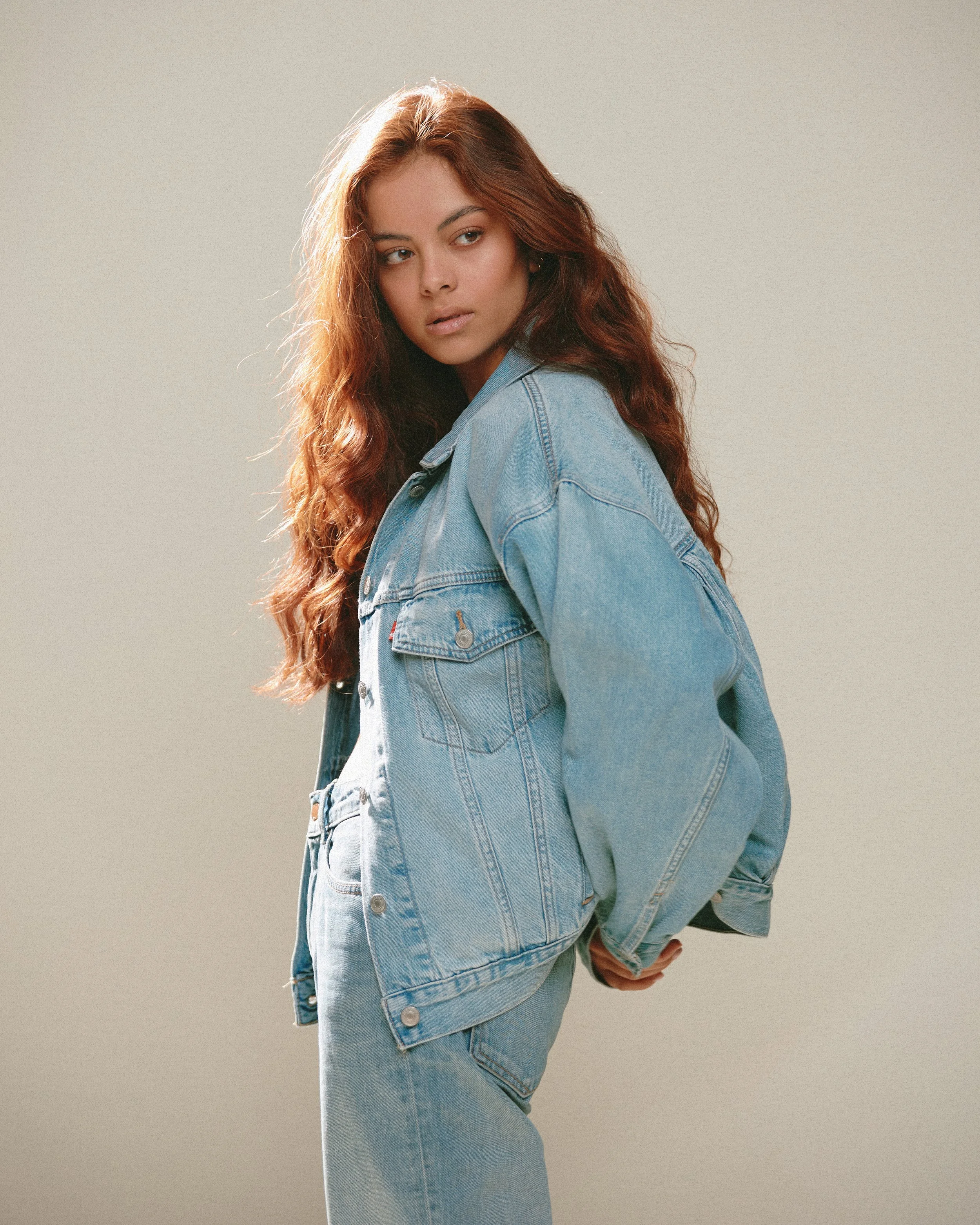 Young woman with long red hair wearing a light blue denim jacket and jeans, posing against a neutral background.