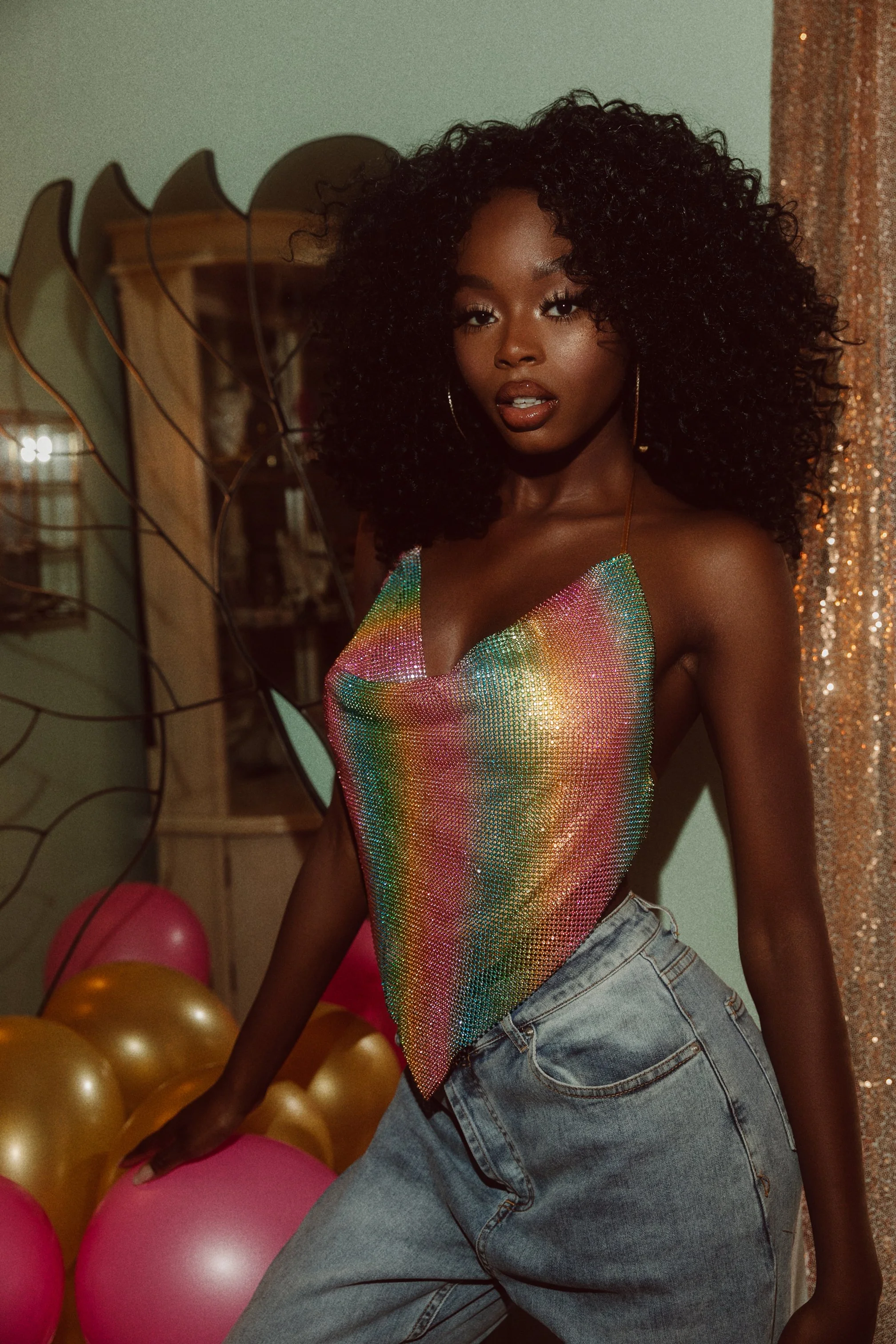 A woman with curly black hair posing indoors, wearing a multicolored, shiny, sleeveless top and light blue jeans, with colorful balloons in the background.