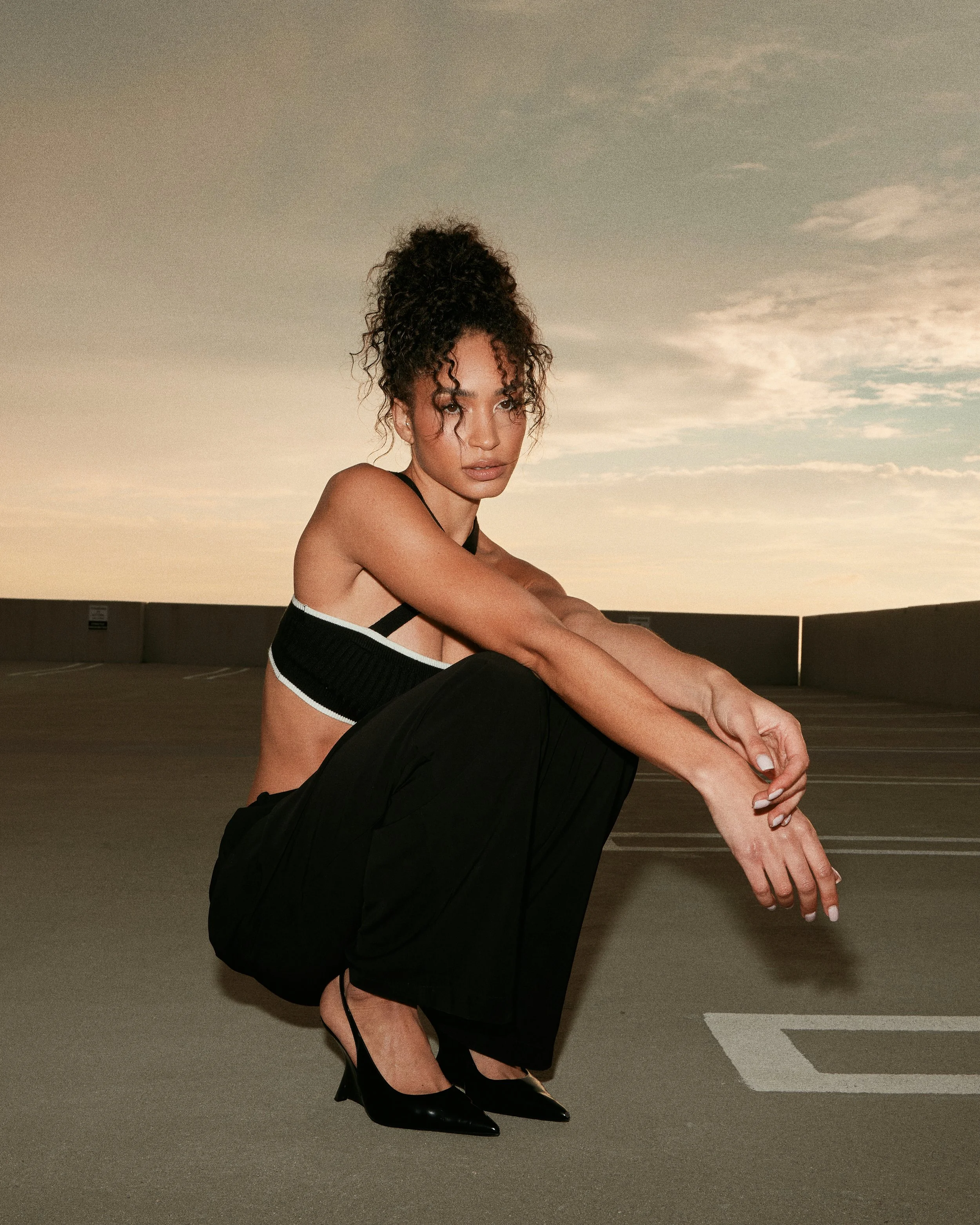 A woman squatting in a parking lot at sunset, wearing a black sports bra and black wide-leg pants, with black heels and curly hair up.
