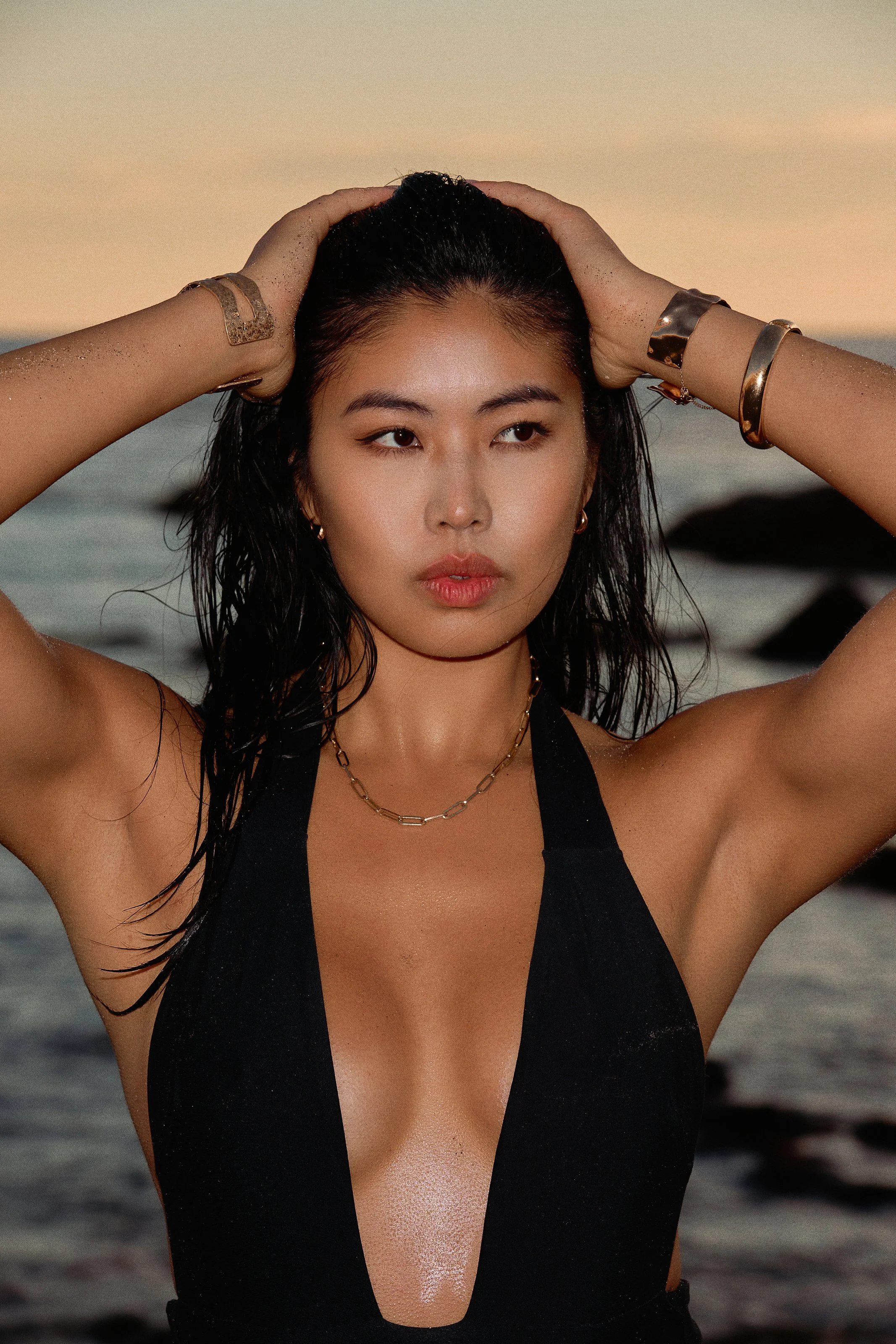 A woman with wet hair poses at the beach during sunset, wearing a black swimsuit and jewelry.