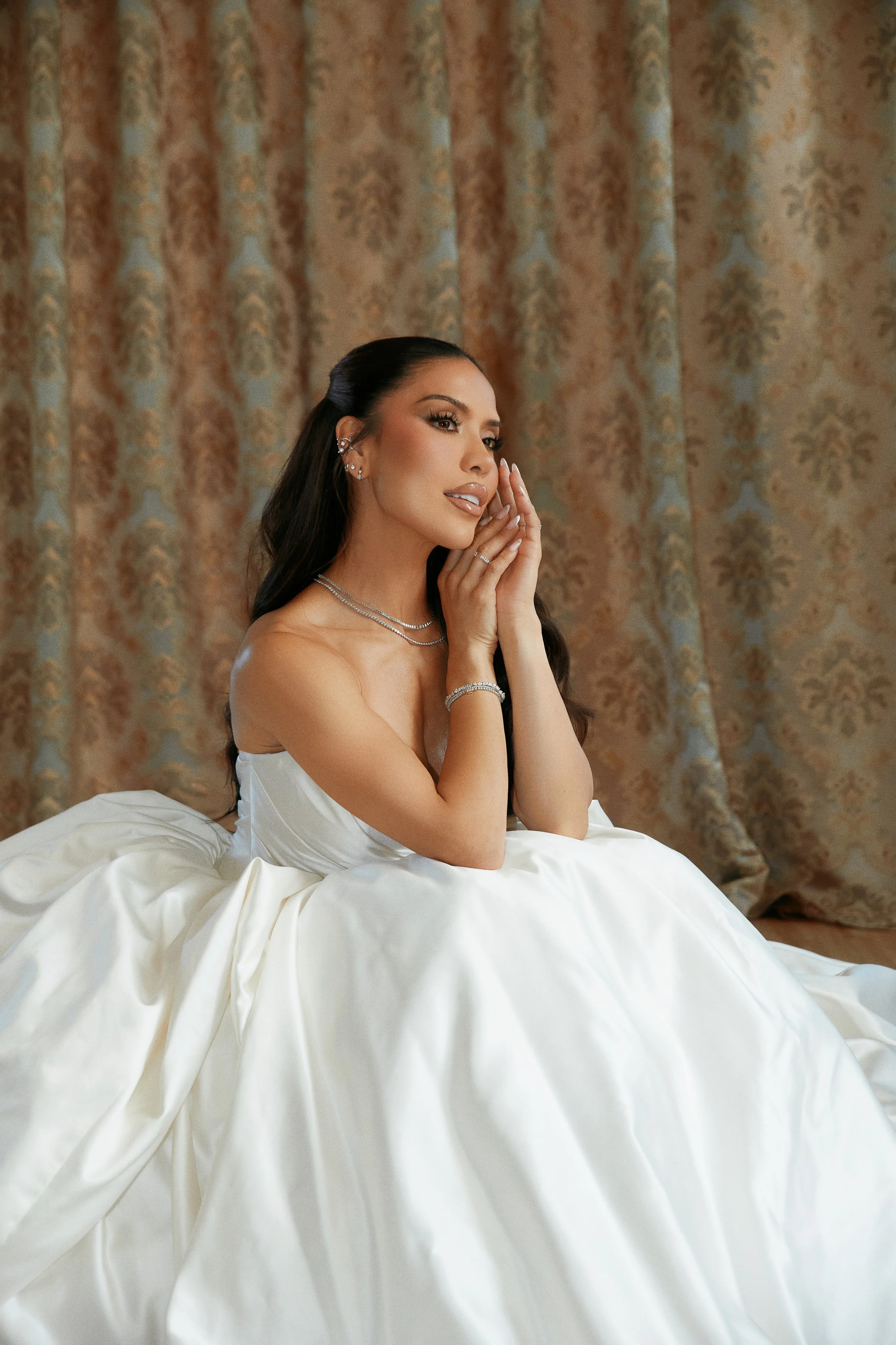 A woman in a white wedding dress sitting in front of a patterned curtain, with her hands gently touching her face.