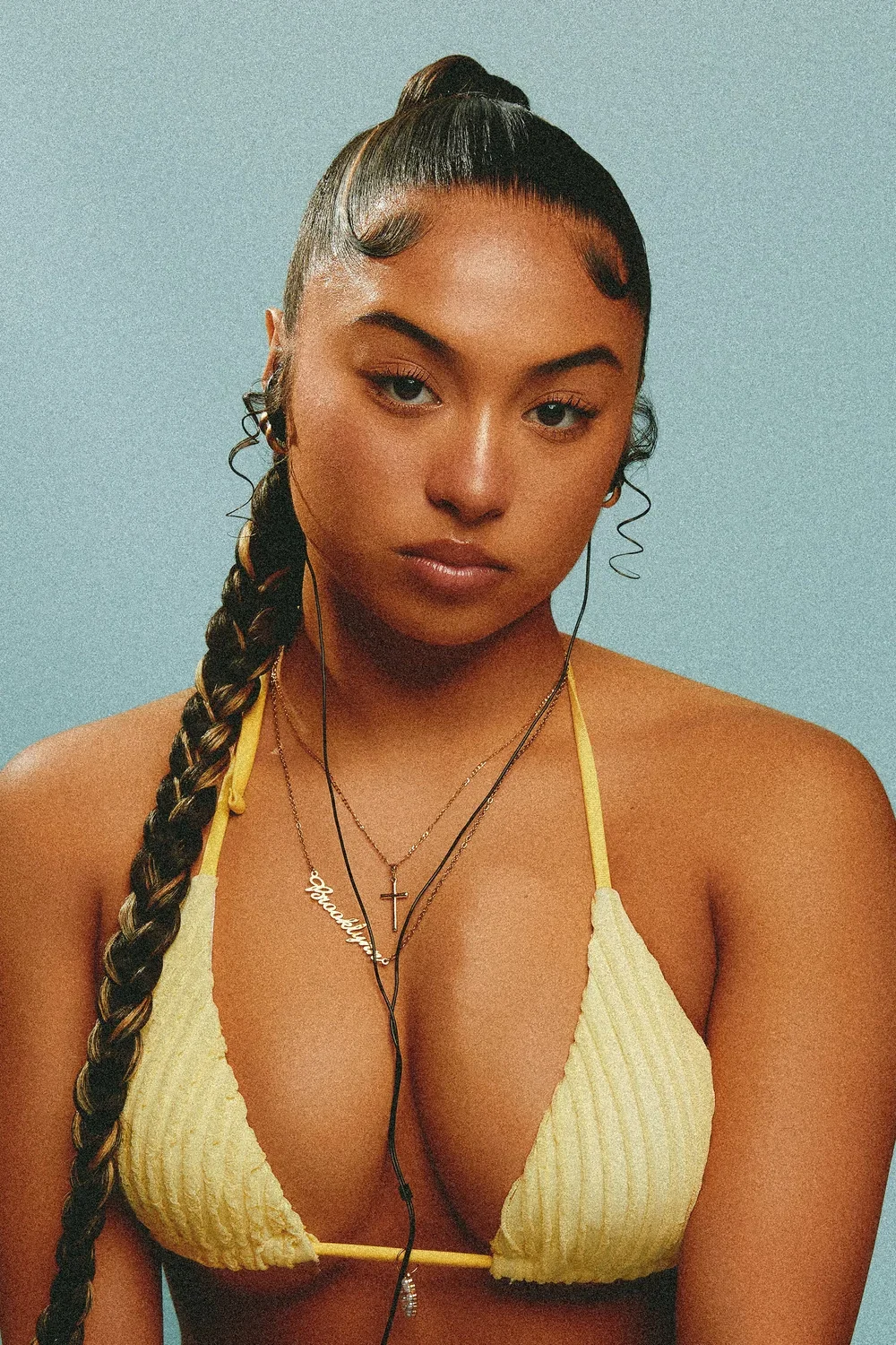 A young woman with braided hair, wearing a yellow bikini and layered necklaces, looking into the camera against a plain background.