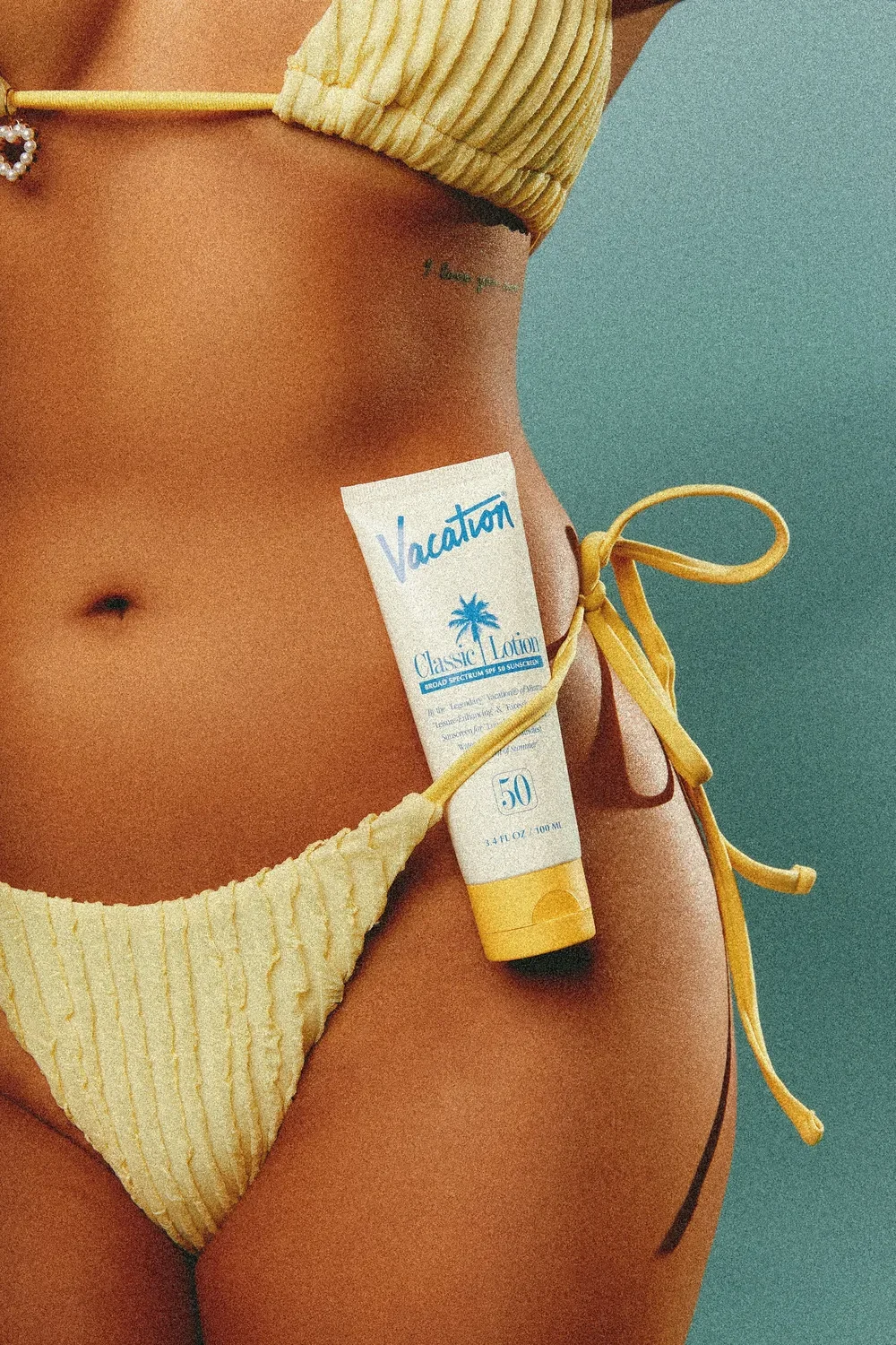 Close-up of a woman's torso in a yellow bikini with string ties, holding a tube of sunscreen labeled 'Vacation Classic Lotion SPF 50' against her side.