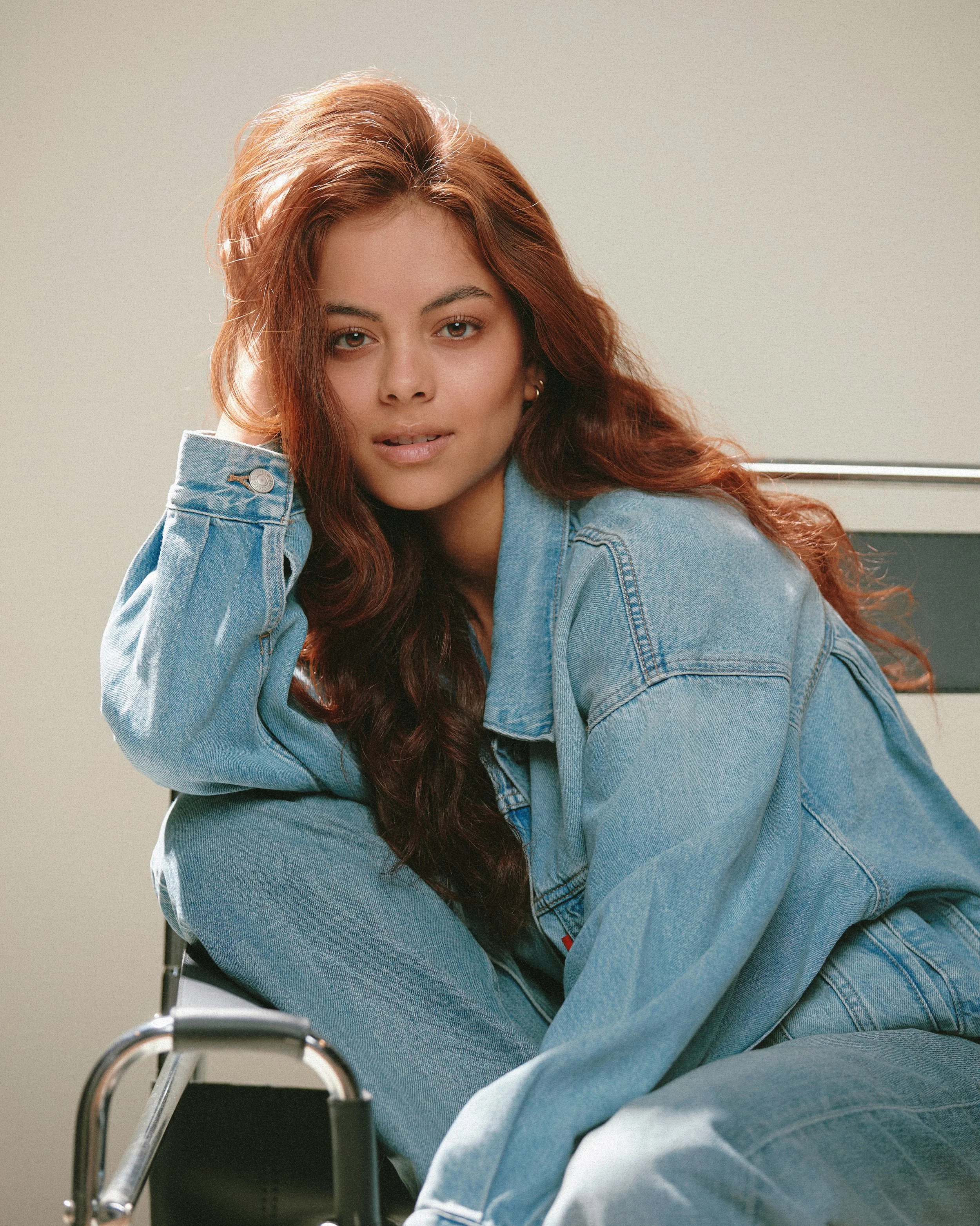 Young woman with long, wavy red hair sitting on a chair, wearing a denim jacket and jeans, looking at the camera with a relaxed expression.