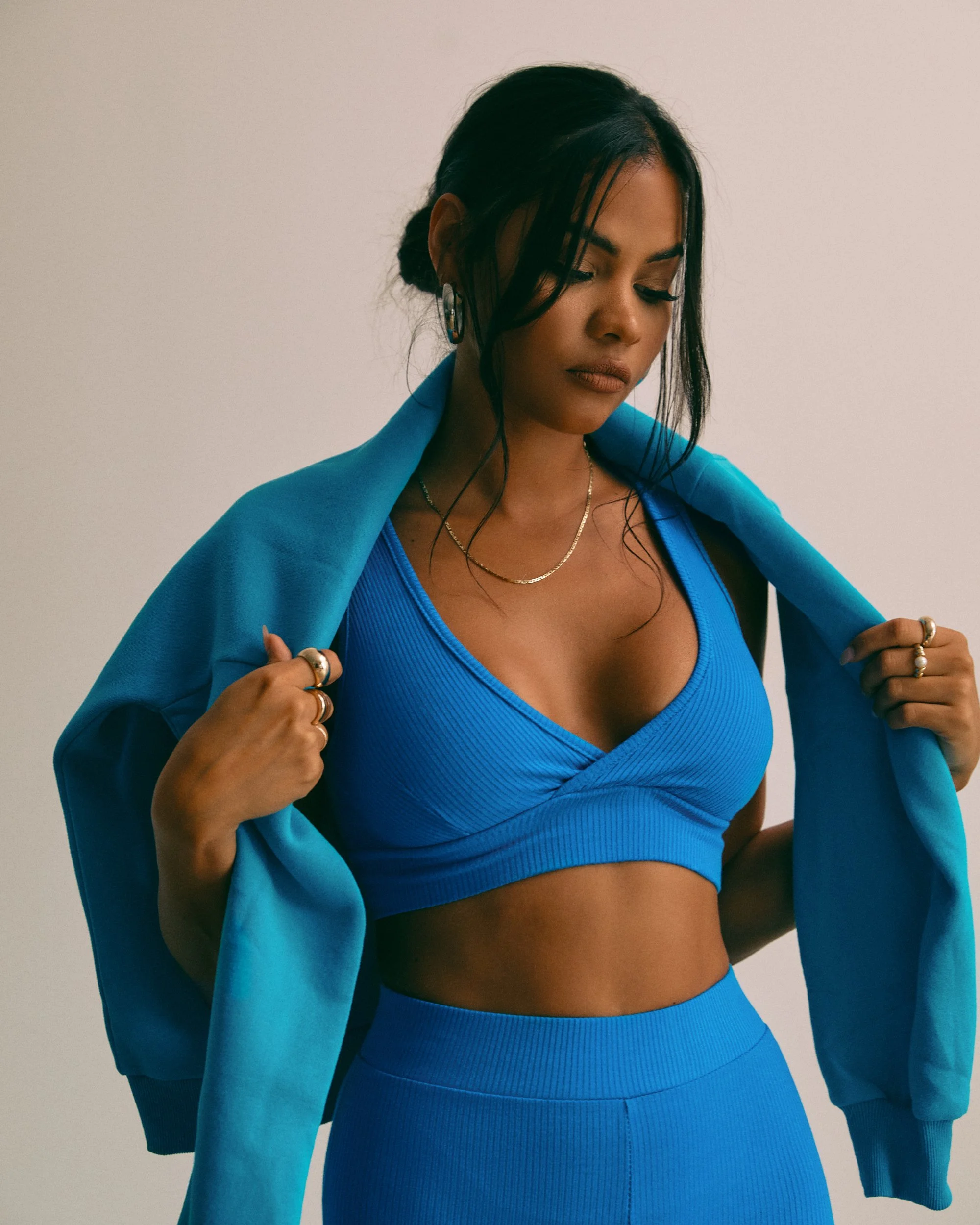 Woman in a blue sports bra and matching leggings, holding a blue jacket over her shoulders, with long black hair tied back, wearing jewelry, looking down.