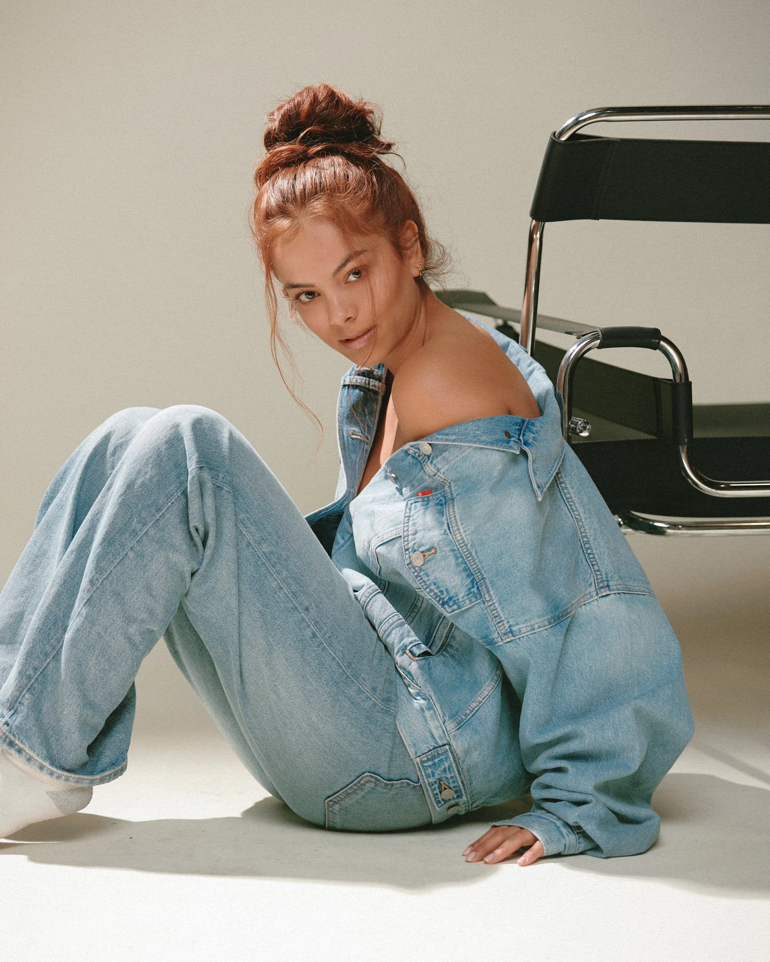 A young woman with red hair in a high bun sitting on the floor, wearing a denim jacket off her shoulders, jeans, and white shoes, next to a black and chrome chair.