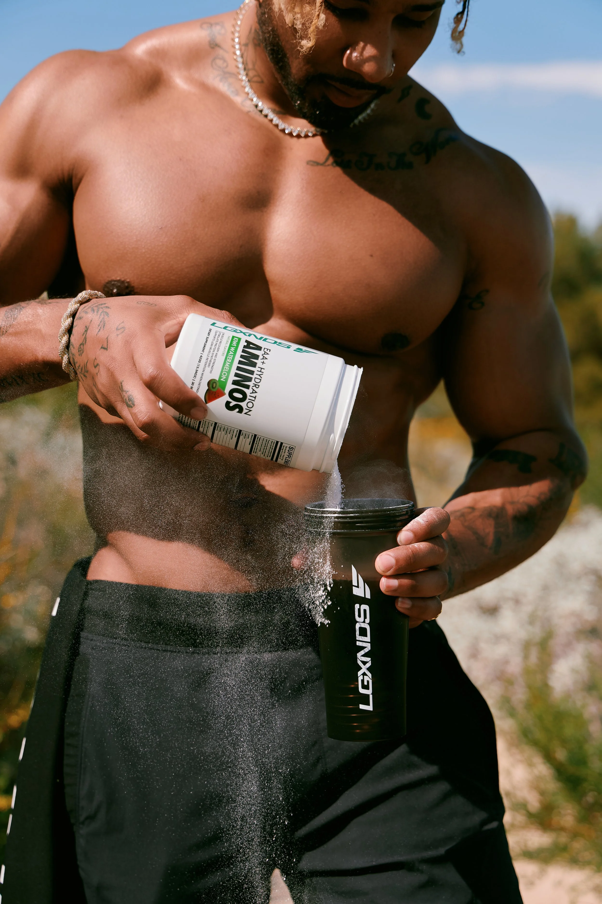 A shirtless man with tattoos and a necklace, outdoors on a sunny day, is pouring powdered supplements from a white container into a black shaker bottle with the logo "LEXSND".