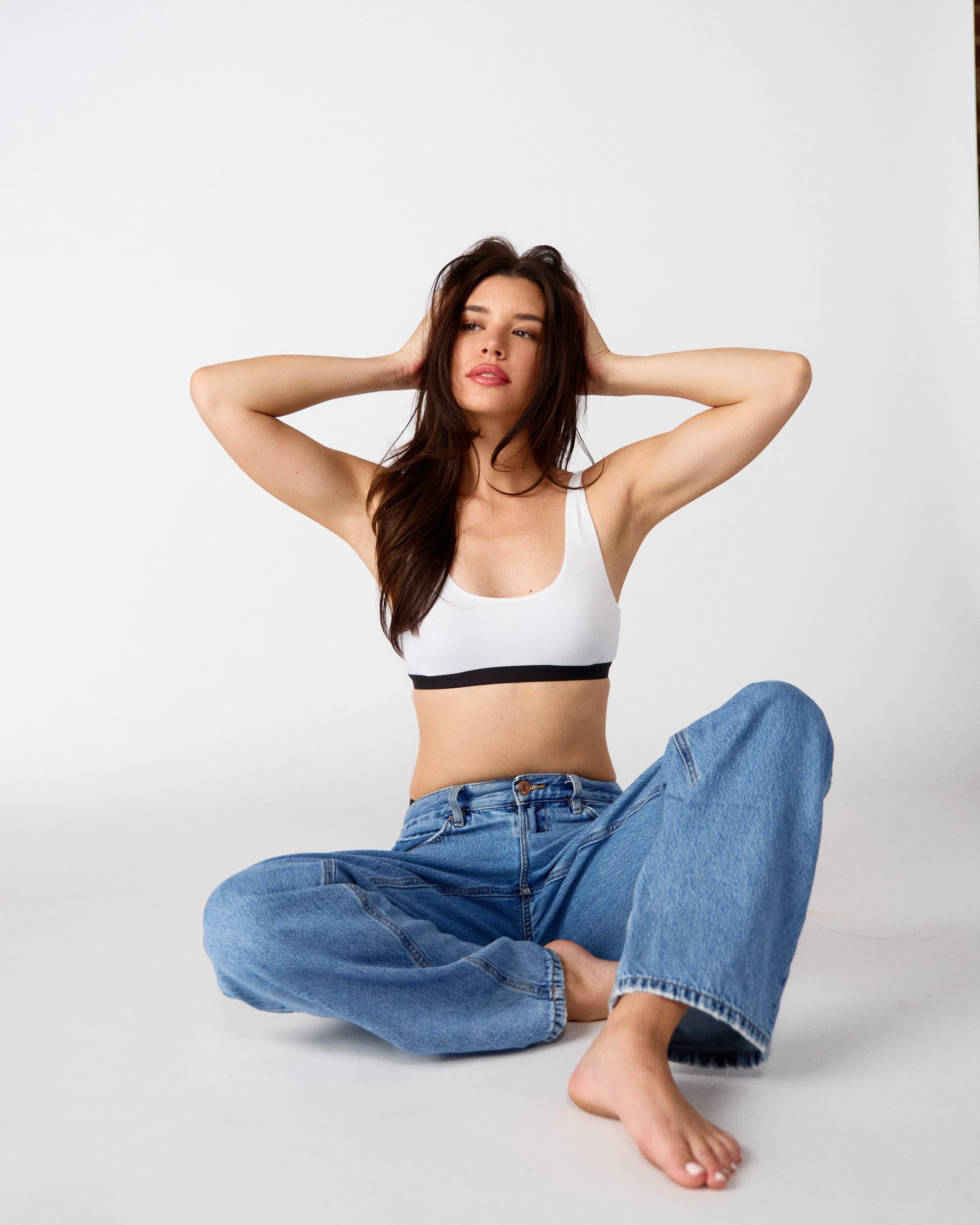 A young woman with long brown hair sitting cross-legged on the floor with her hands behind her head, wearing a white sports bra and loose blue jeans, against a plain white background.