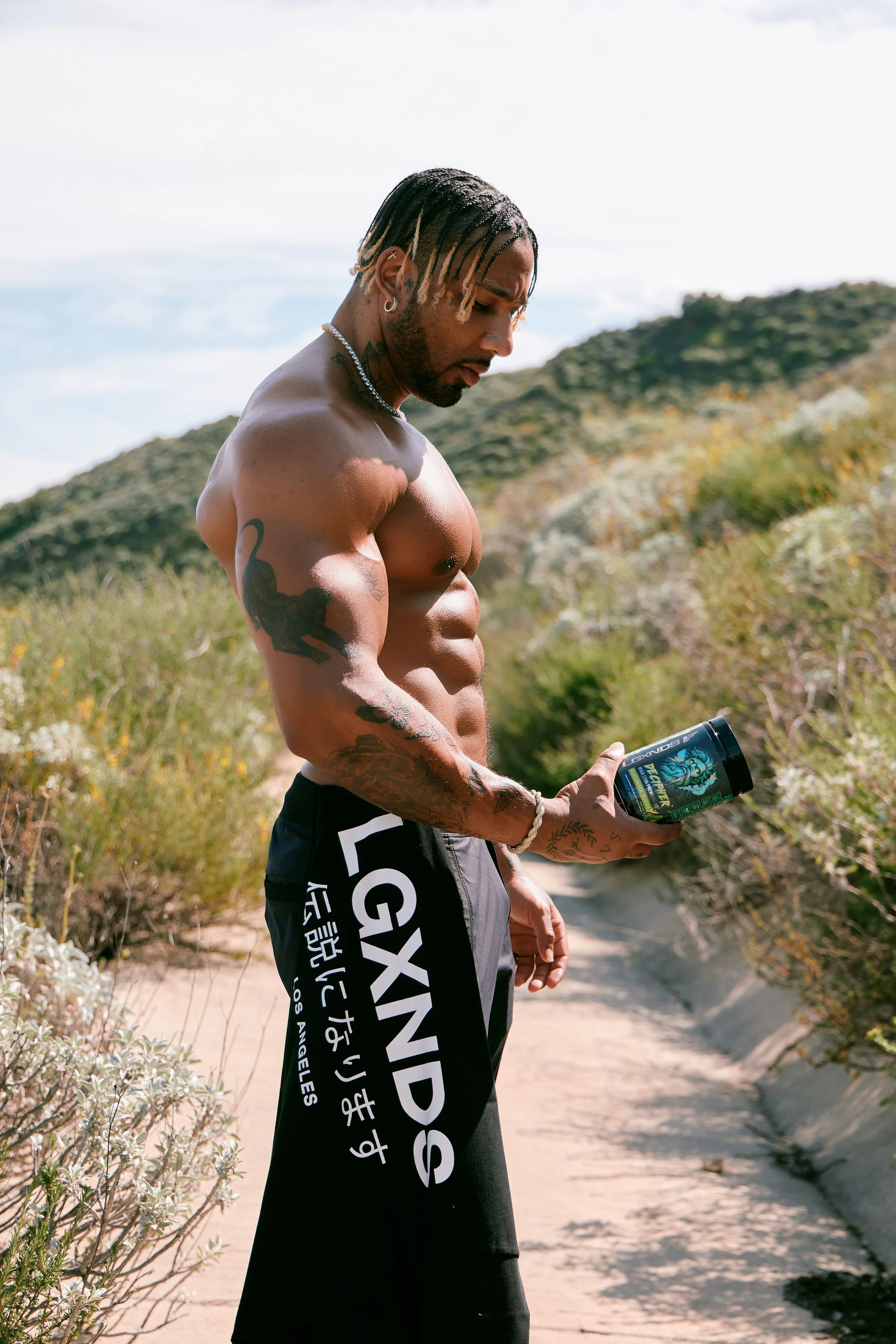 A muscular man with tattoos, wearing black shorts with 'LAXNPS' and Japanese characters, standing outdoors on a trail in a hilly, wooded area, looking at a container of dietary supplement.