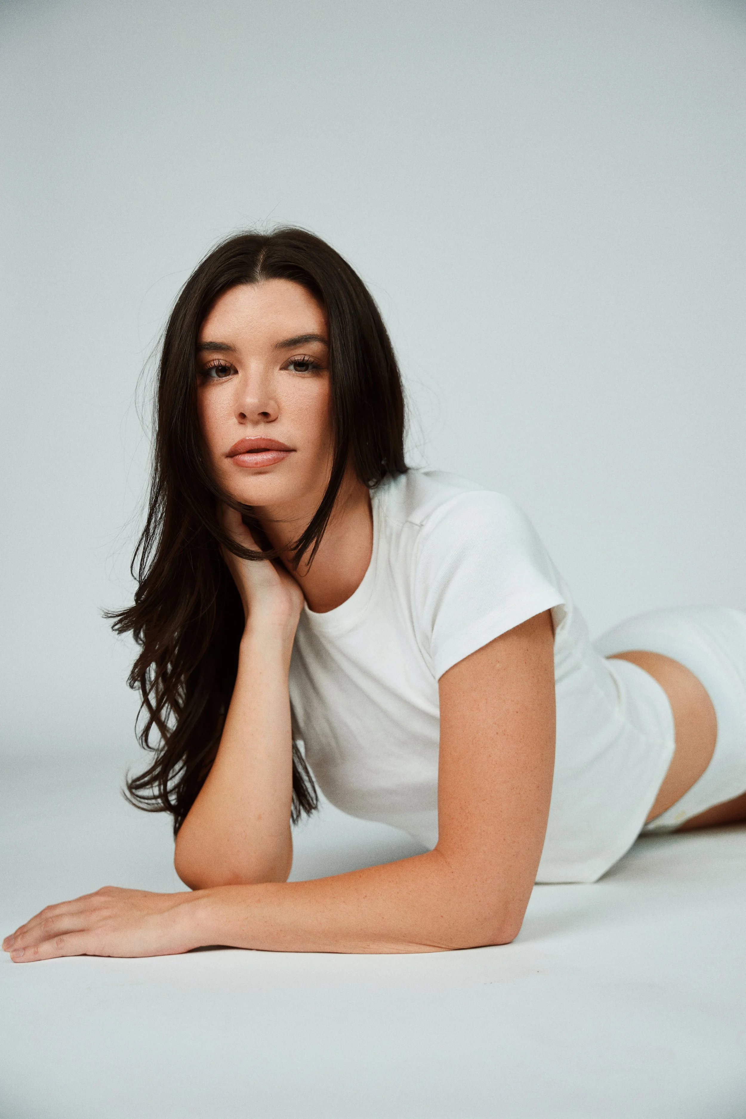 A young woman with dark brown hair in a white t-shirt and white shorts lying on her side on a plain white background, resting her head on her hand and looking at the camera.