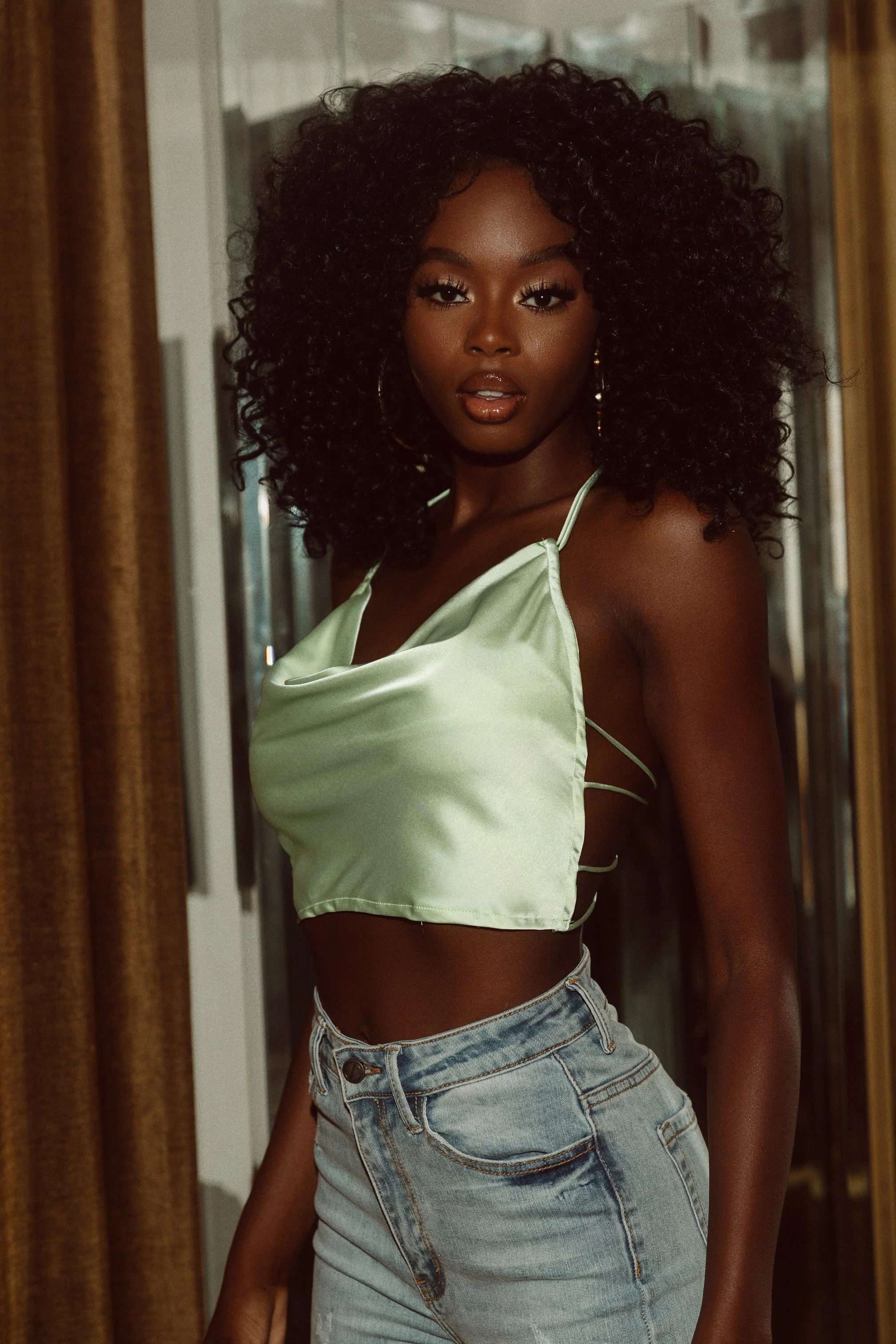 A woman with dark skin and curly black hair posing indoors, wearing a shiny green crop top and high-waisted blue jeans.