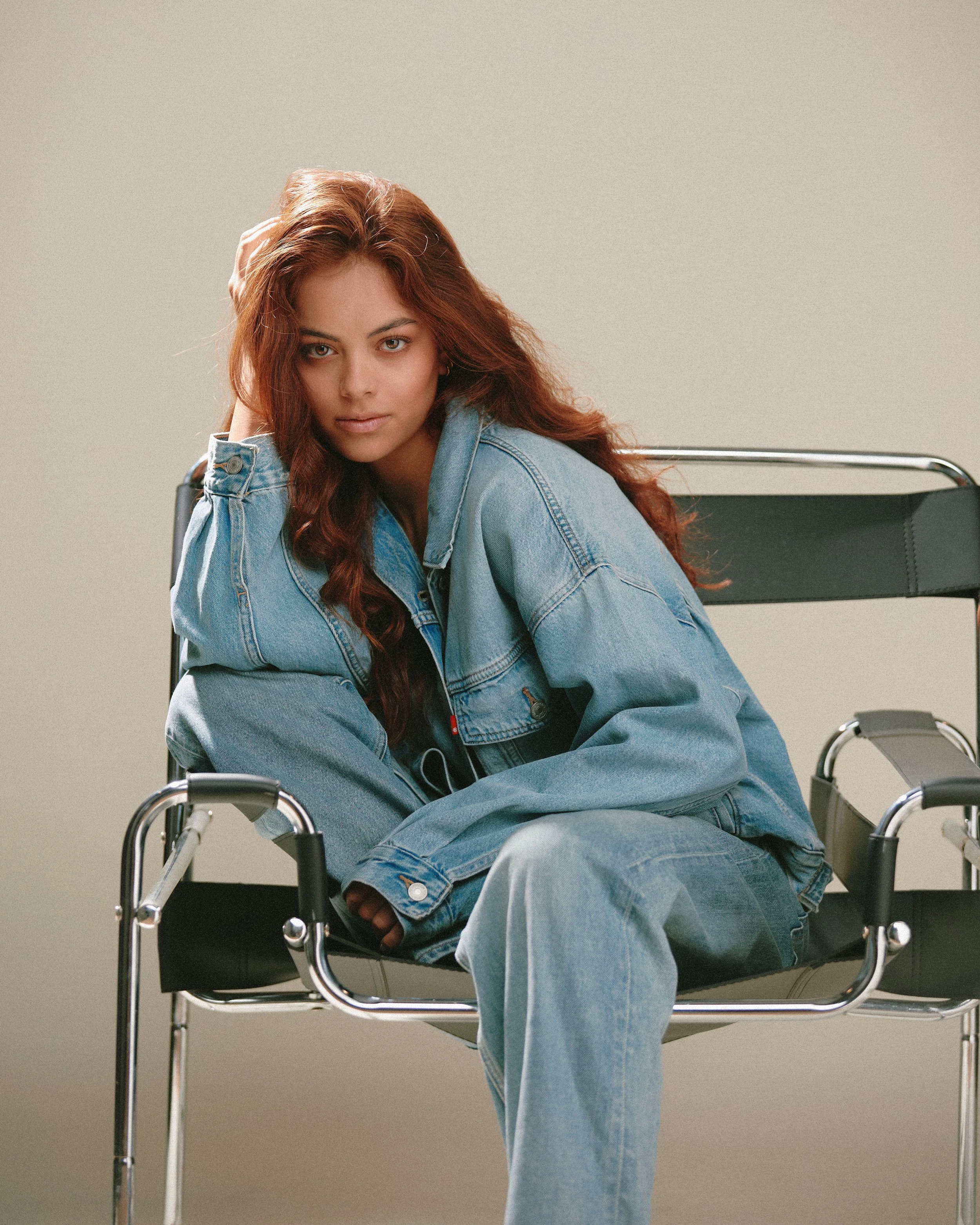A young woman with long, wavy auburn hair sitting on a modern black lounge chair with chrome frame, wearing a blue denim jacket and jeans, looking at the camera with a serious expression, against a plain beige background.