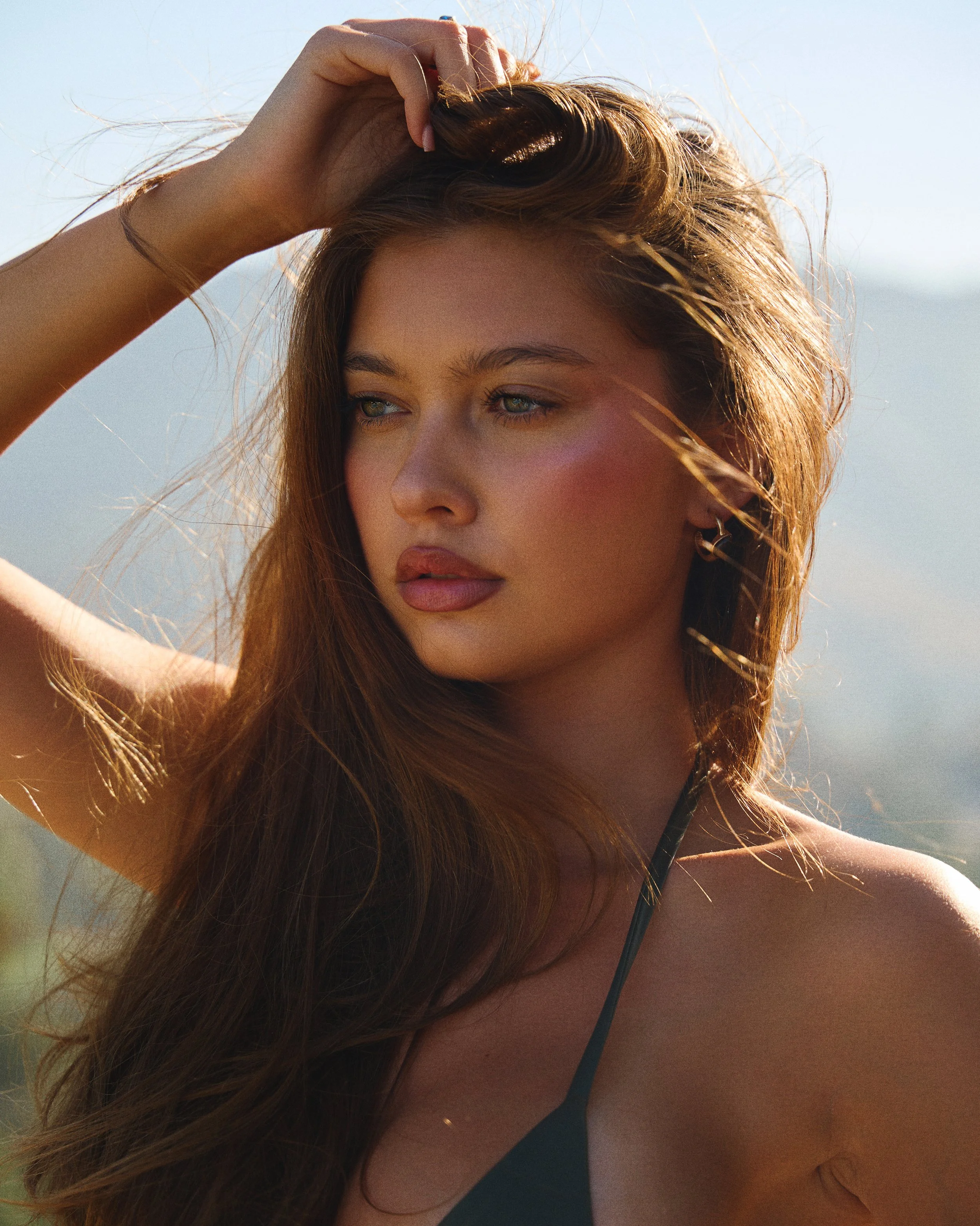 Close-up of a woman with long brown hair and blue eyes, wearing a black top, outdoors during daytime, with an out-of-focus mountain or hill in the background.