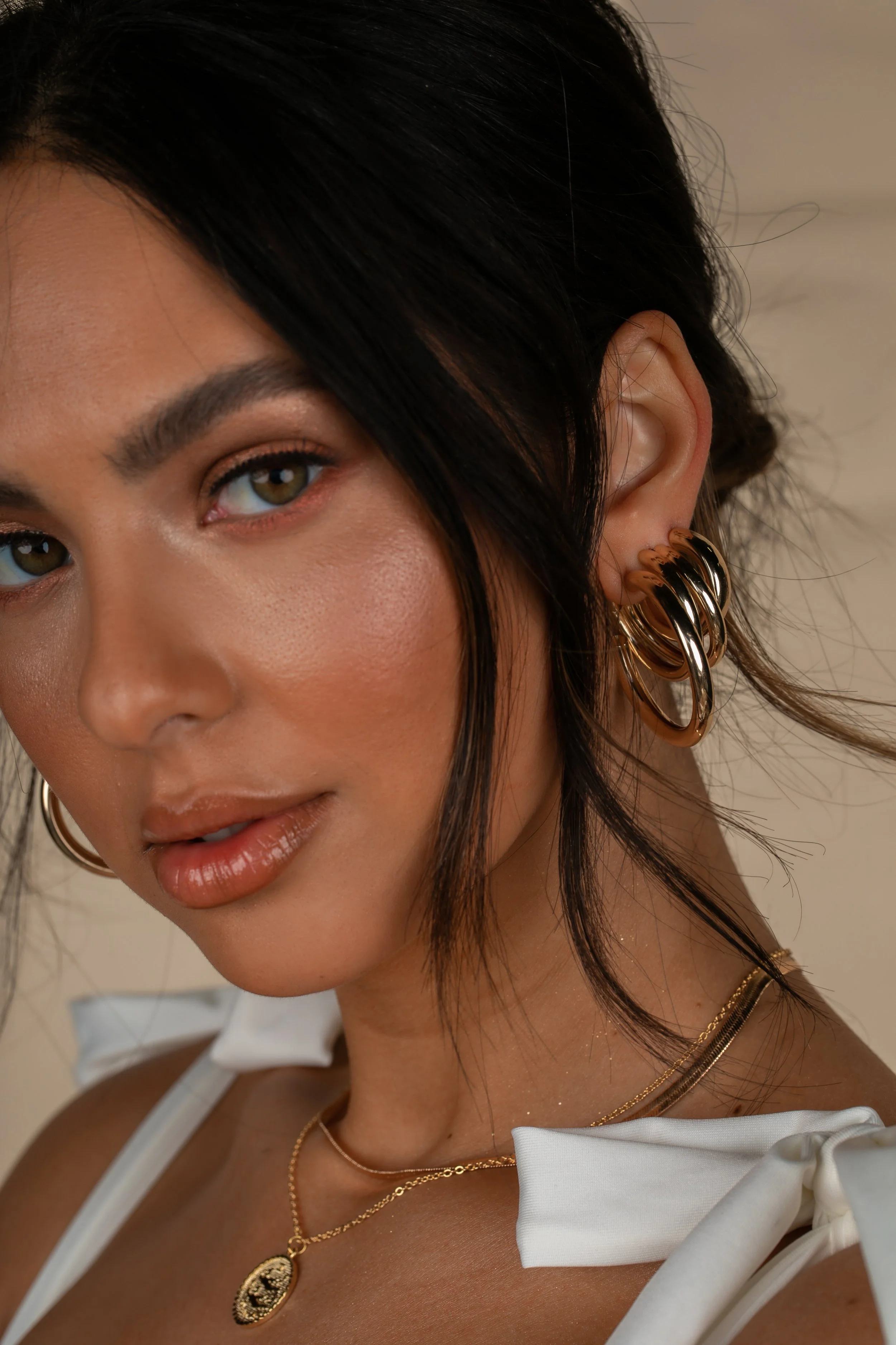 Close-up of a woman with dark hair, wearing multiple gold hoop earrings, layered gold necklaces, and a white top with bow details.