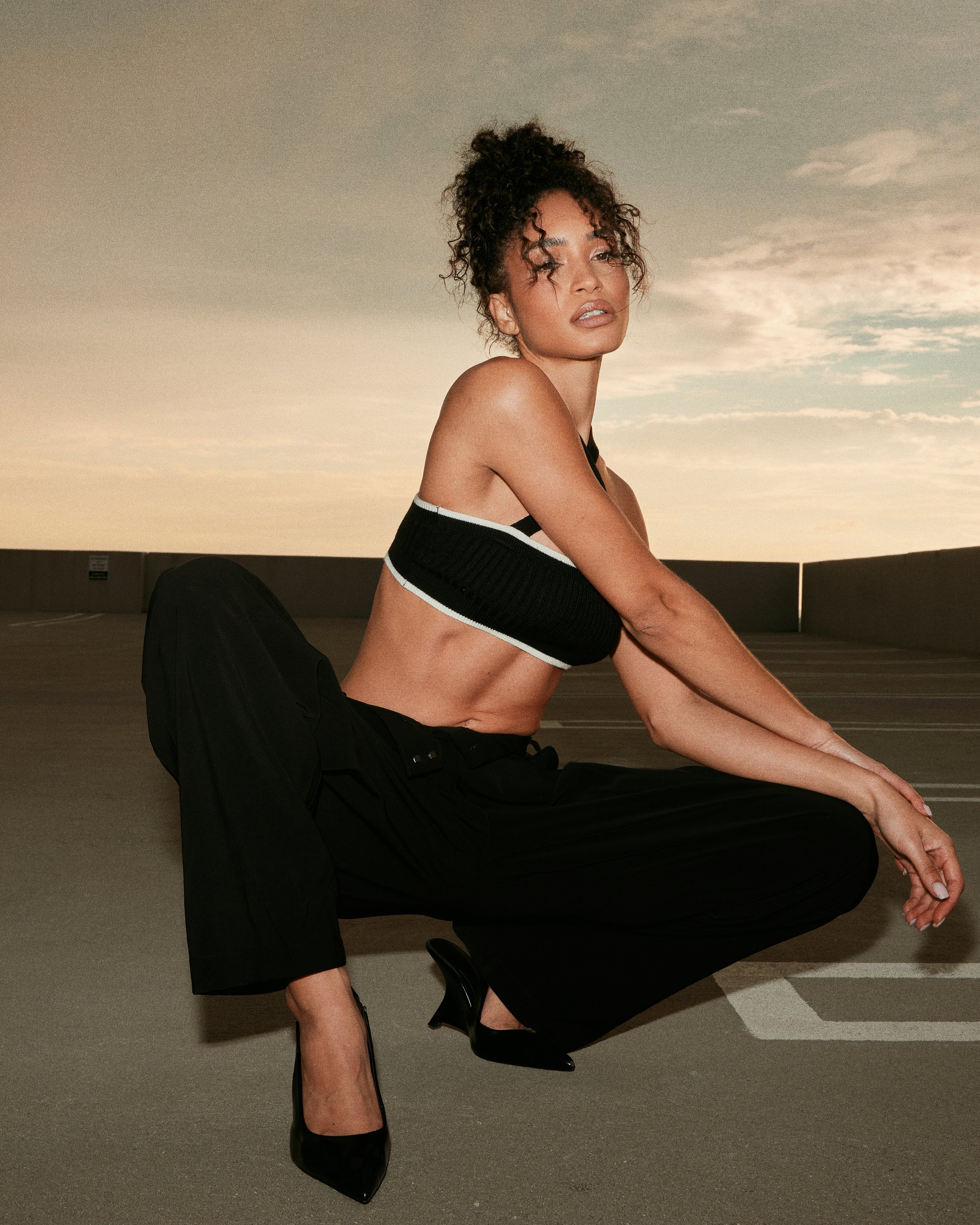 A woman wearing a black sleeveless top, black pants, and black high heels in a squatting pose on a rooftop parking lot during sunset.
