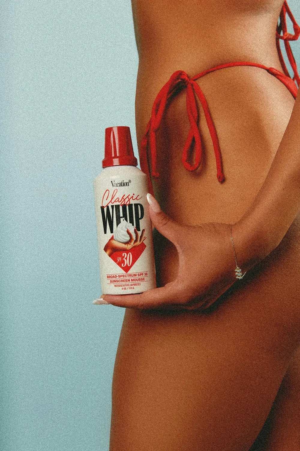 Person in red bikini holding a bottle of sunscreen labeled 'Vacation Classic Whip SPF 30' against a light blue background.