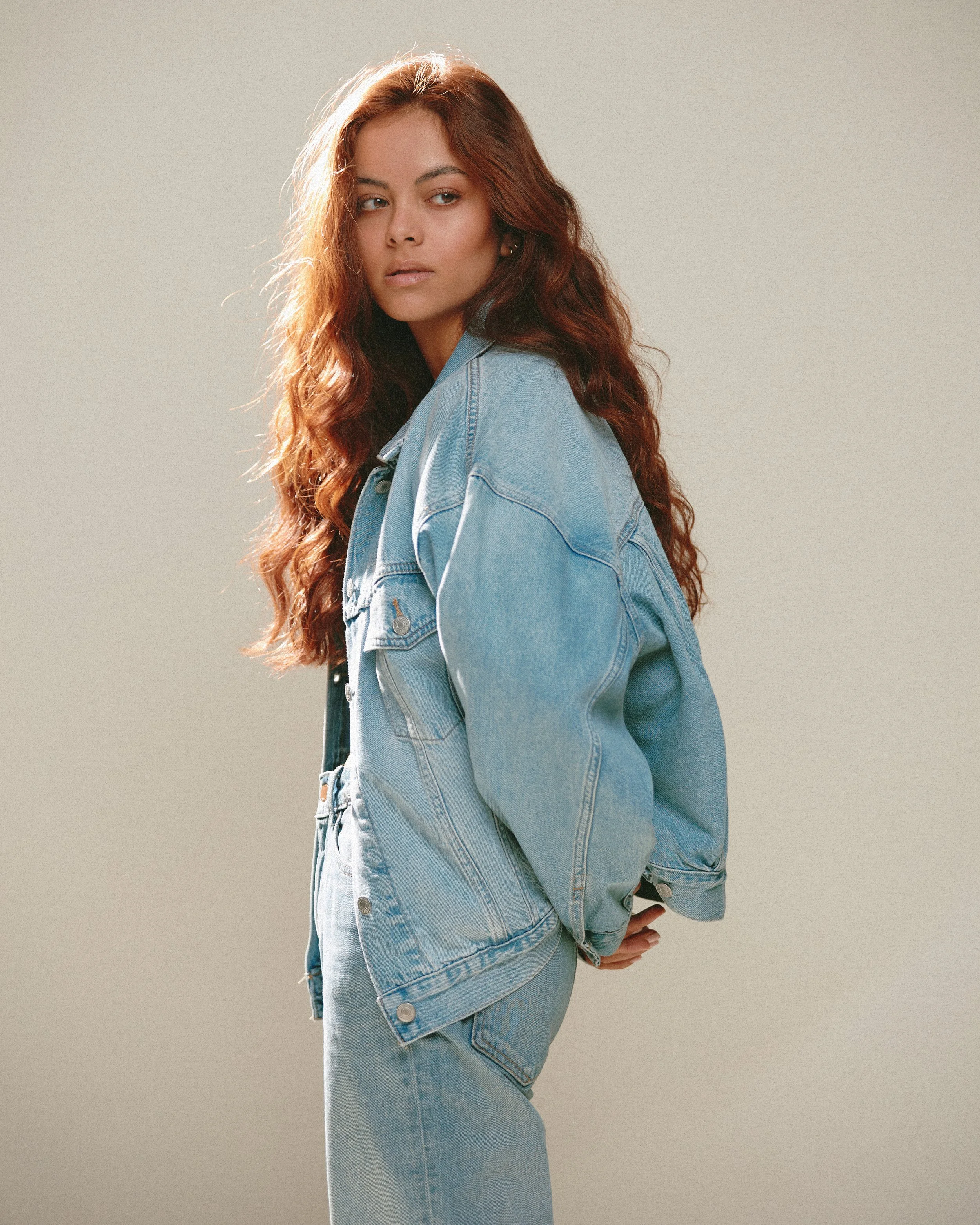 Woman with long, wavy red hair wearing a light denim jacket and jeans posing against a plain background.