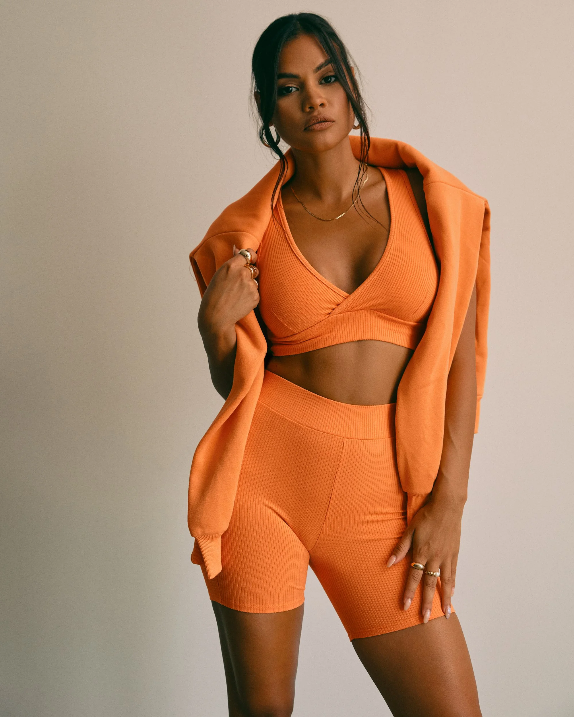 A woman in orange athletic wear with a matching jacket draped over her shoulders, standing against a plain background.
