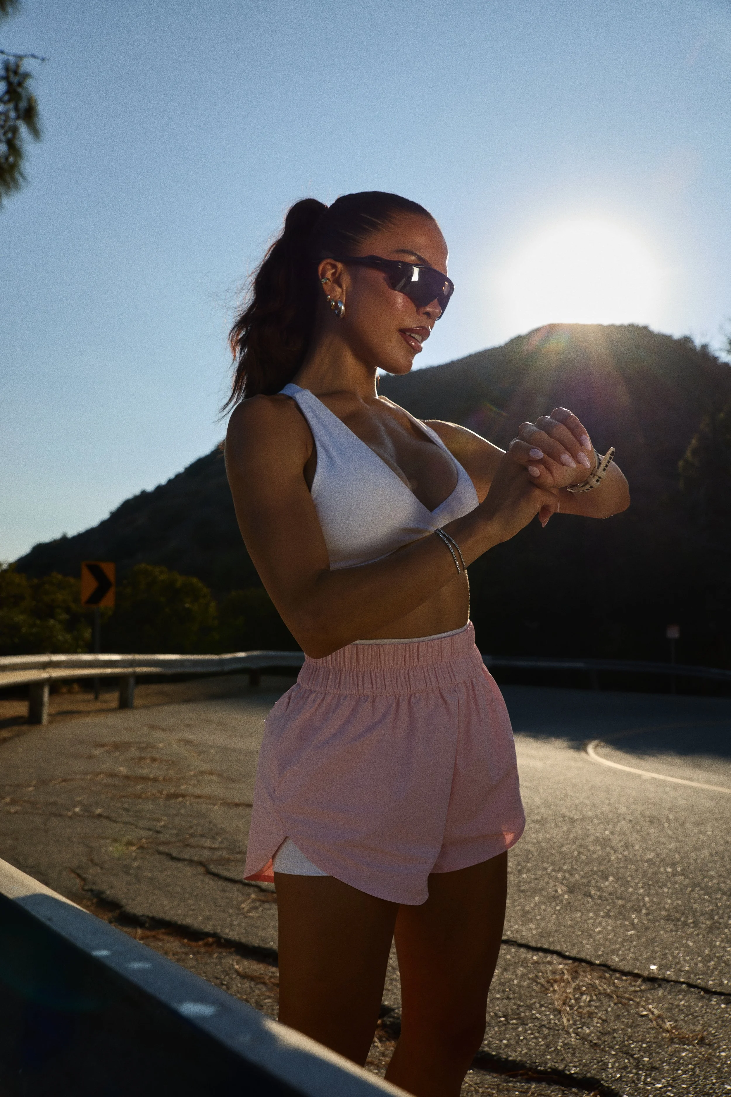 A woman wearing sunglasses and athletic attire checking her watch outdoors with a mountain and the sun in the background.