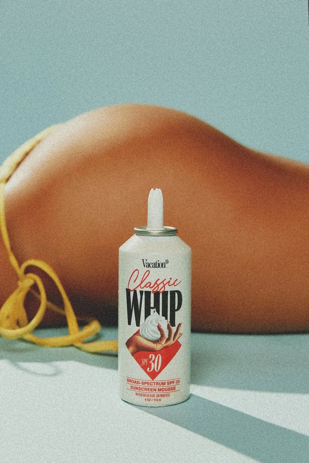 A can of broad-spectrum SPF 30 sunscreen mousse labeled 'Vacation Classic Whip' with a yellow ribbon and a hand holding the mousse, set against a blurred background of a beach setting with a tan-colored surface.