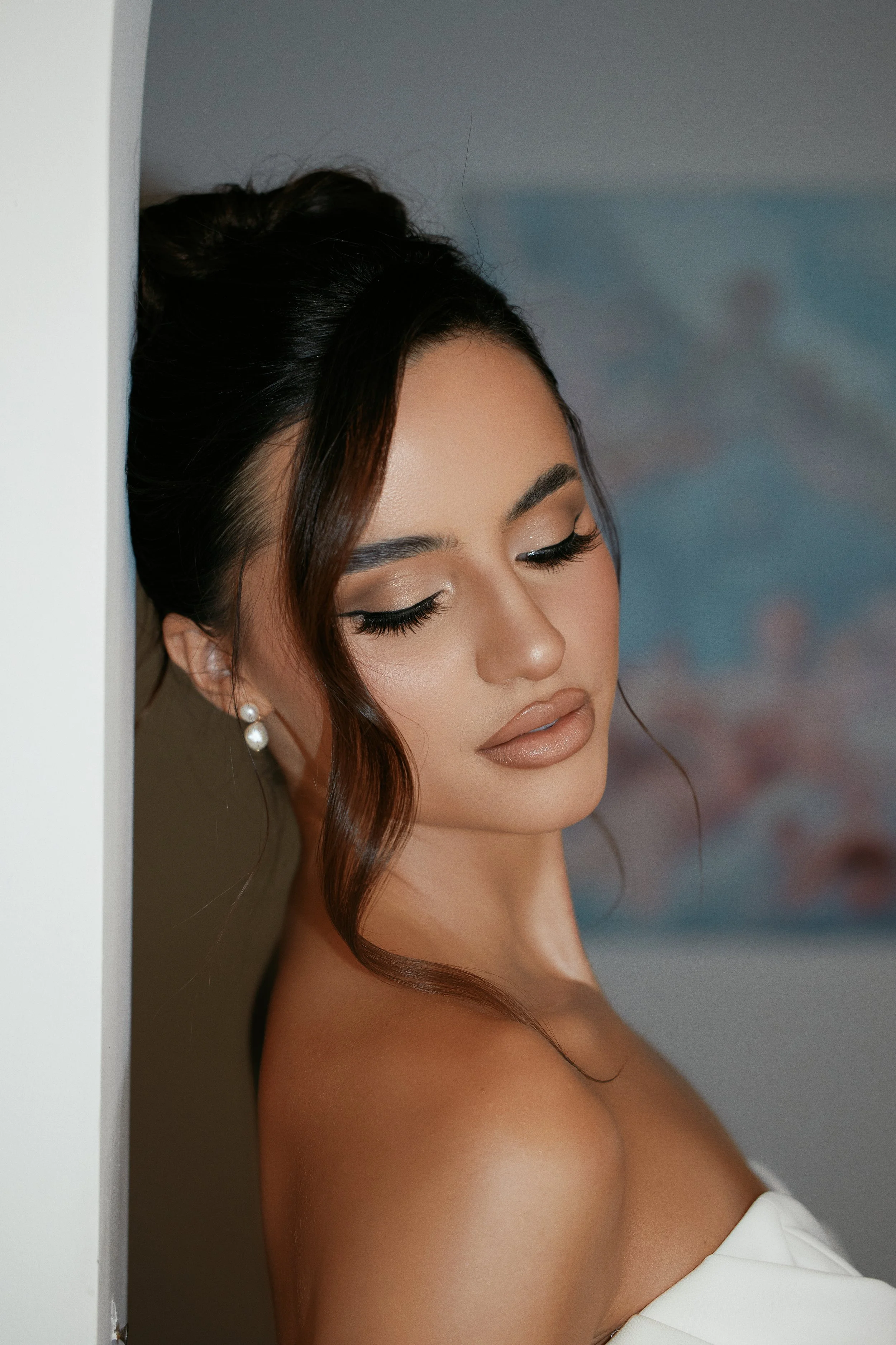 A close-up of a woman with makeup and pearl earrings, with her eyes closed, leaning against a wall.