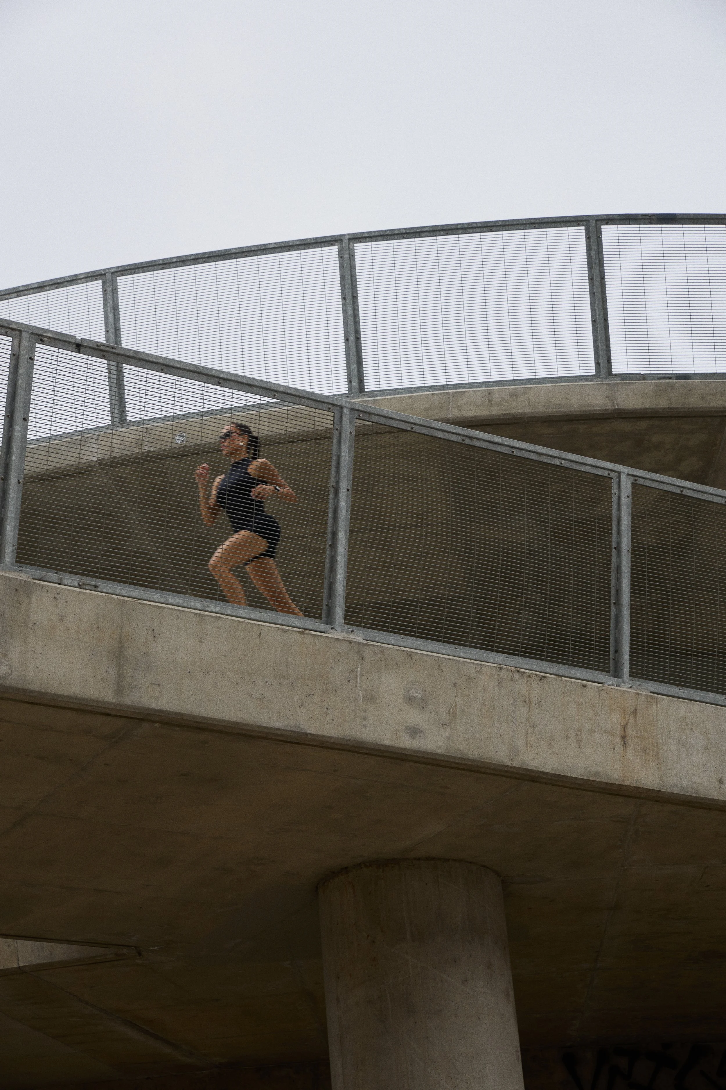 A woman running on an outdoor concrete elevated pathway with metal fencing, under an overcast sky.