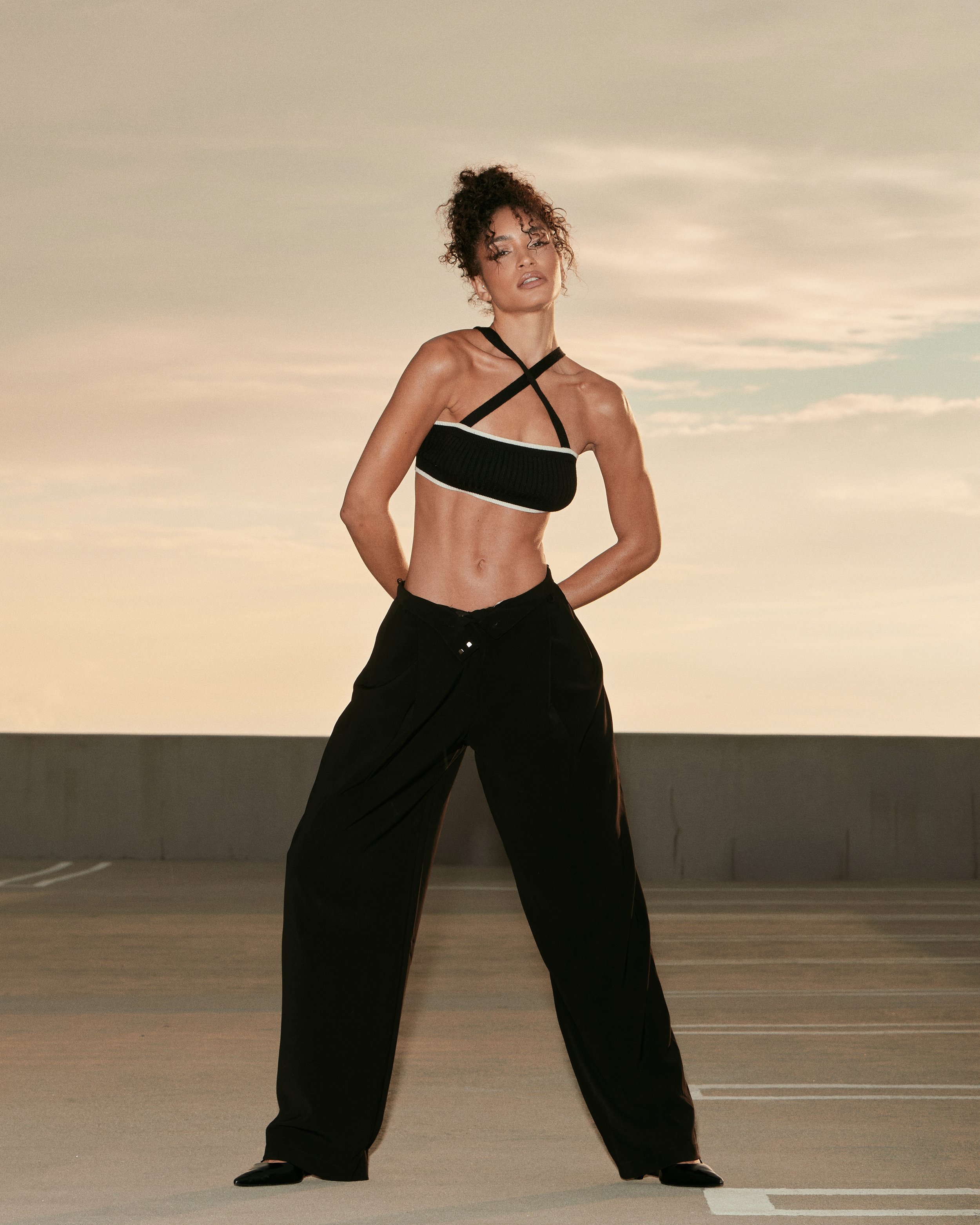 A woman with curly hair standing on a rooftop parking lot during sunset, wearing a black crop top with crisscross straps and loose black pants.