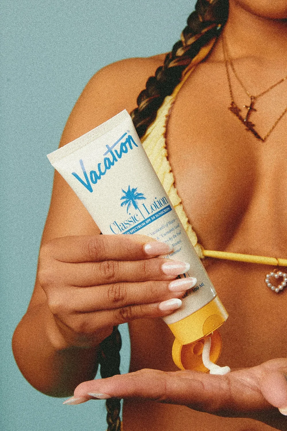 A woman holding a tube of Valatum classic lotion sunscreen with a palm tree logo, wearing layered necklaces, and sporting a braided hairstyle.