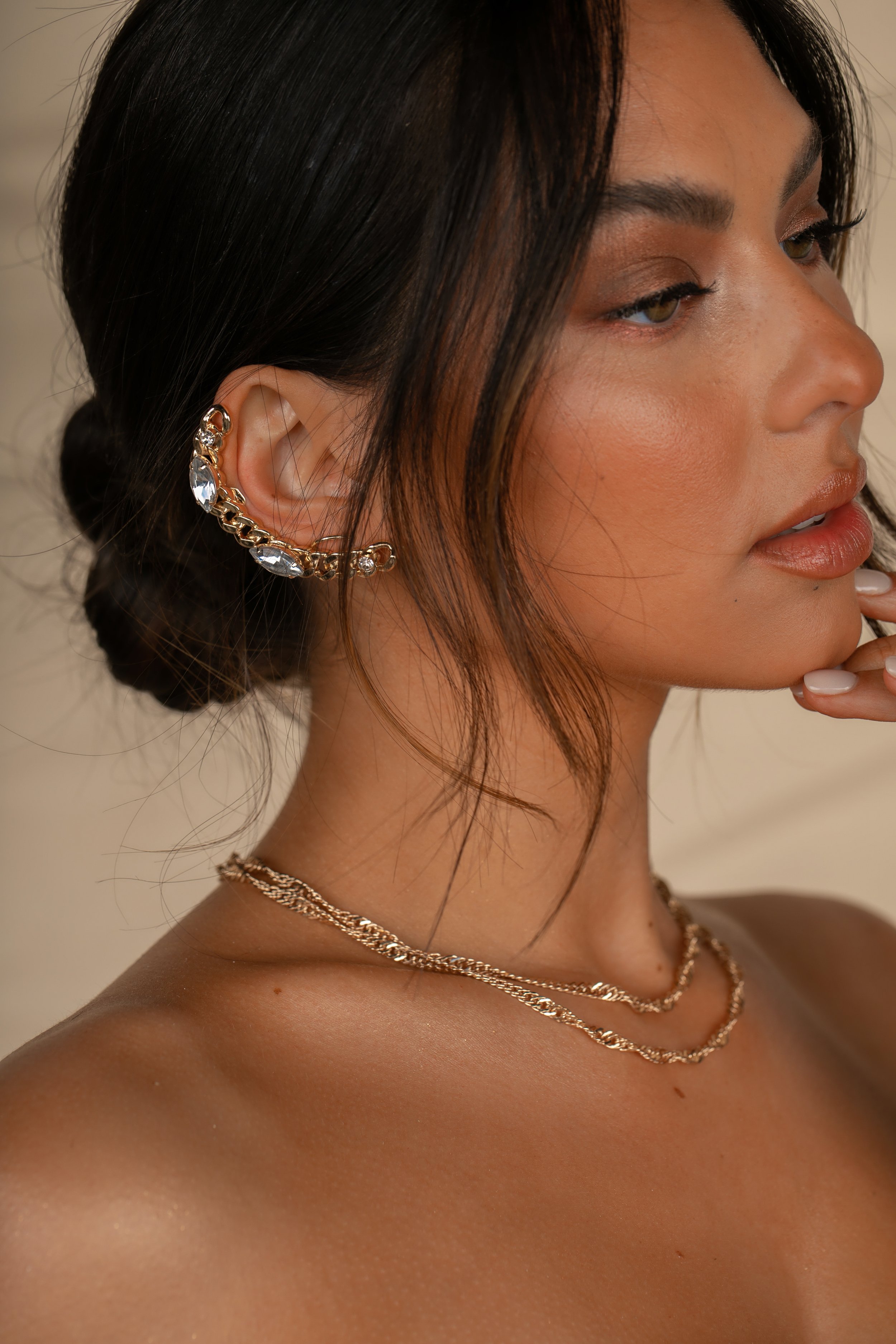 Close-up of a woman's face, showing her dark hair styled in a bun, wearing a gold ear cuff with clear stones and layered gold necklaces.