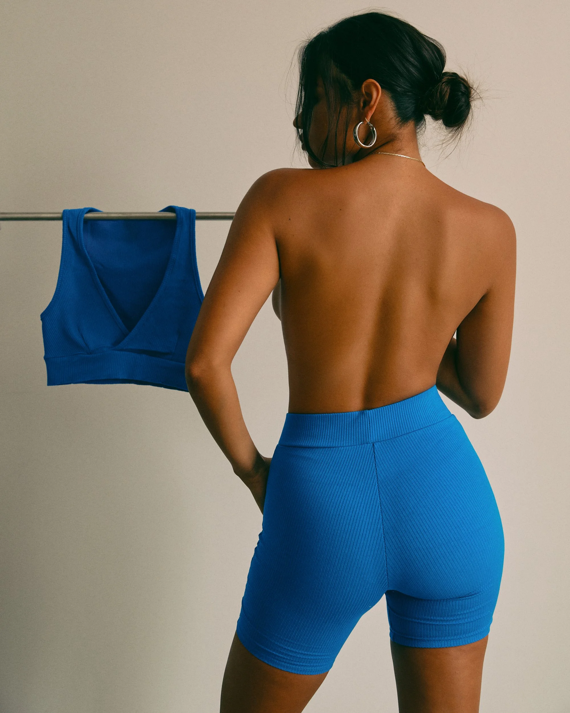 Back view of a woman with dark hair tied in a bun, wearing blue high-waisted shorts, standing next to a clothing rack with a blue sports bra.