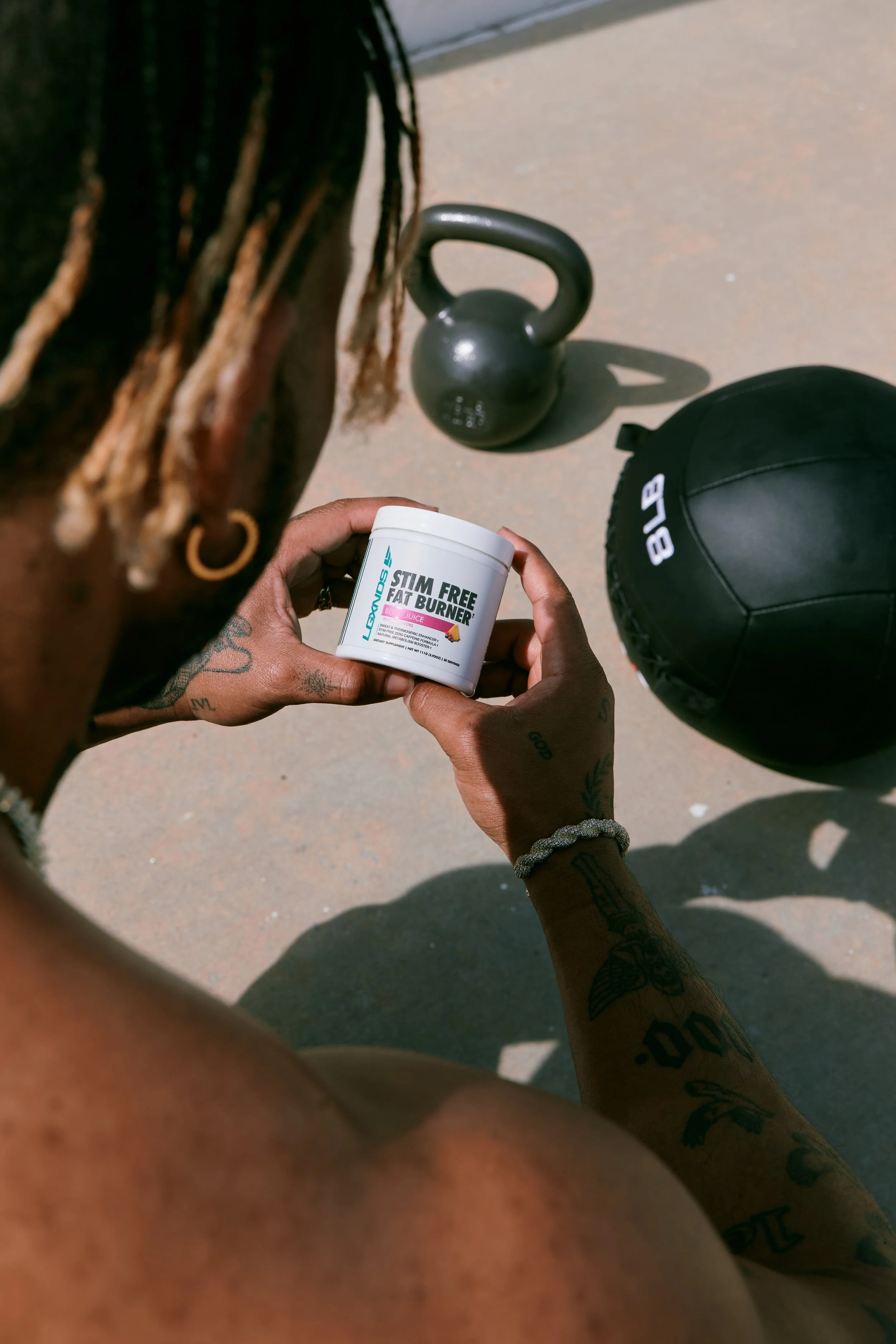 A person with tattoos holding a container of 'Stim Free Fat Burner' supplement, with a kettlebell and a medicine ball on the ground nearby.