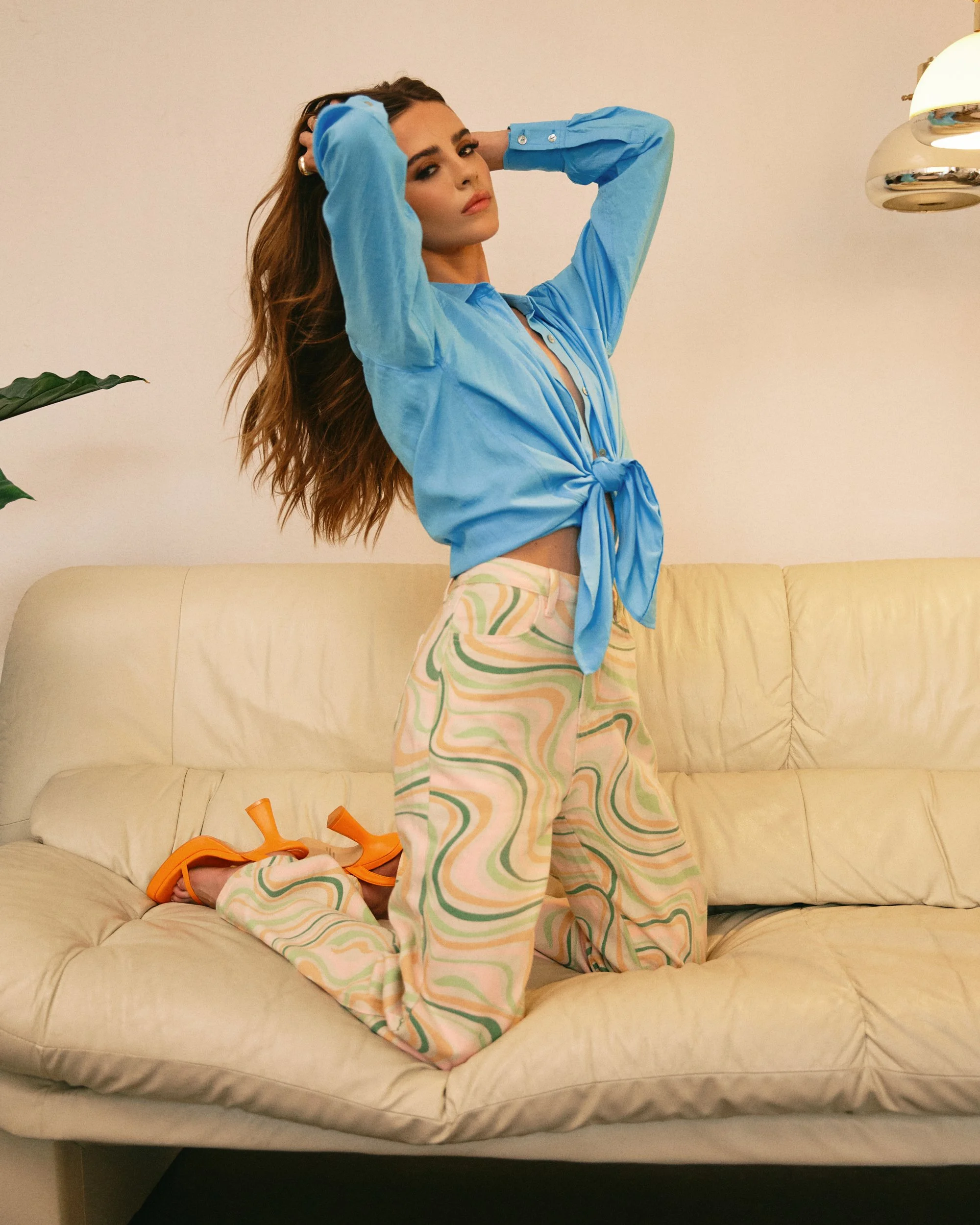A woman with long hair kneels on a cream-colored sofa, wearing a tied-up blue button-up shirt, colorful patterned pants, and orange high-heeled sandals. She has her hands behind her head and looks at the camera.