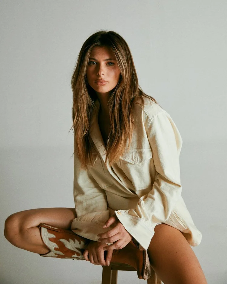 A young woman with long, wavy brown hair sits on a stool against a plain light-colored background. She is wearing a cream-colored oversized jacket and cowboy boots, with one leg bent and resting on the seat of the stool, and her hands clasped in her 