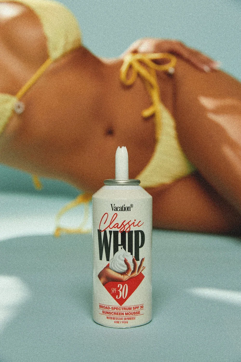 A can of Vacation Classic Whip sunscreen mousse with SPF 30 placed on a flat surface. In the background, a person wearing a yellow bikini top with tied straps is visible, with their hand resting on their shoulder.