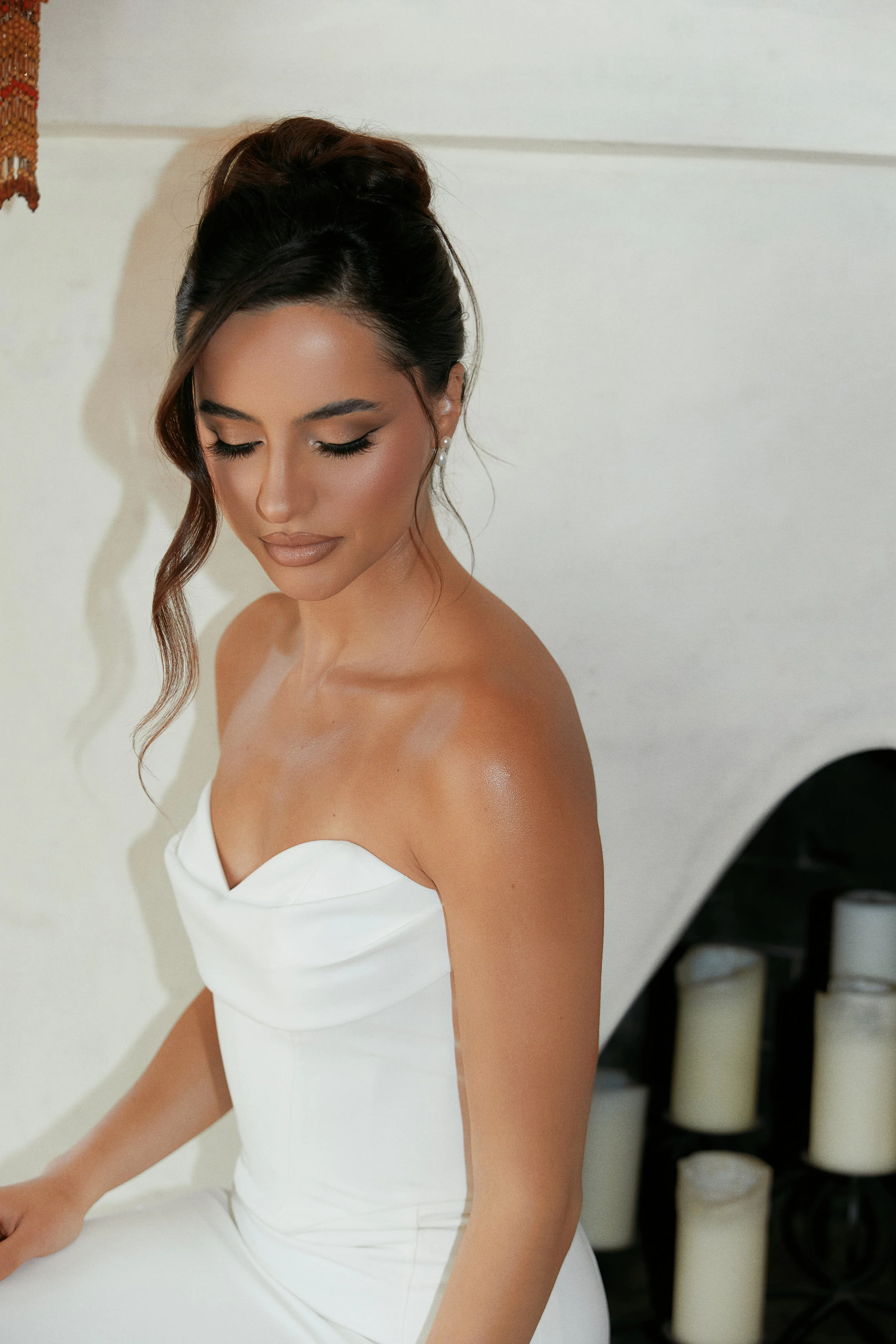 A woman with dark hair styled in an elegant updo, wearing a strapless white dress with a sweetheart neckline, sitting in front of a white background with a few candles in the background.