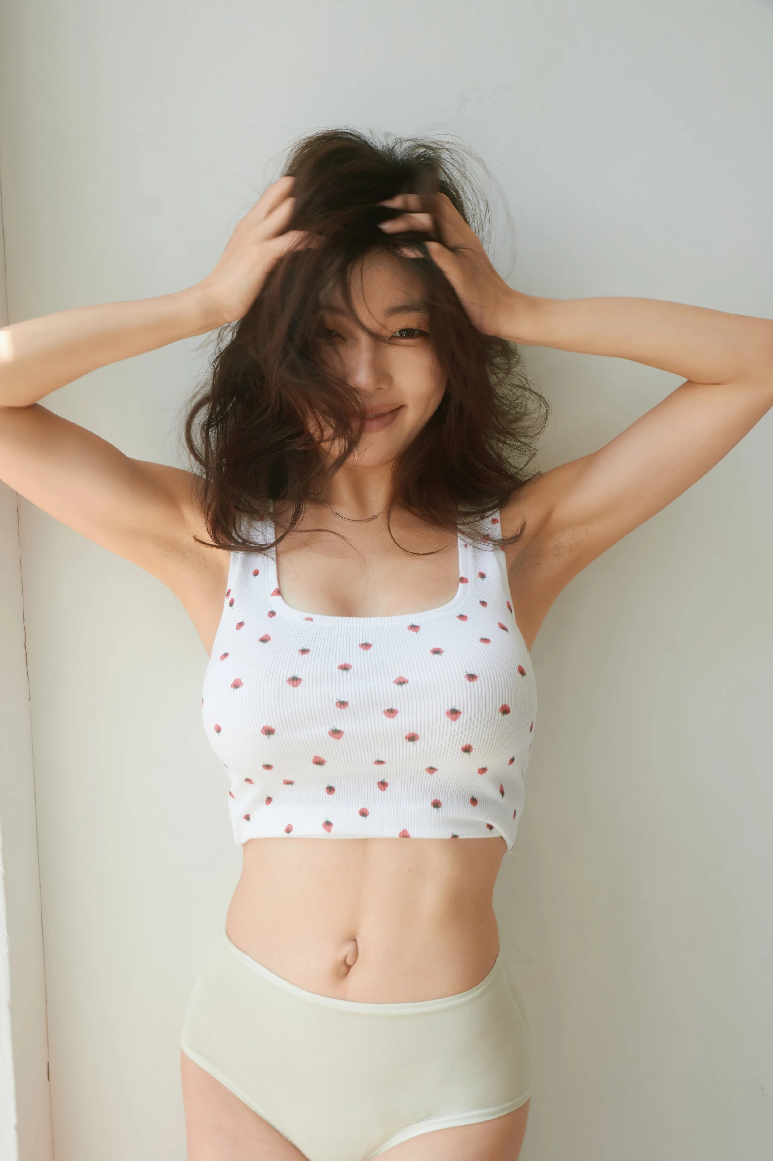 A woman with wavy hair, smiling and touching her hair, wearing a white tank top with a strawberry pattern and beige underwear, standing against a light-colored wall.