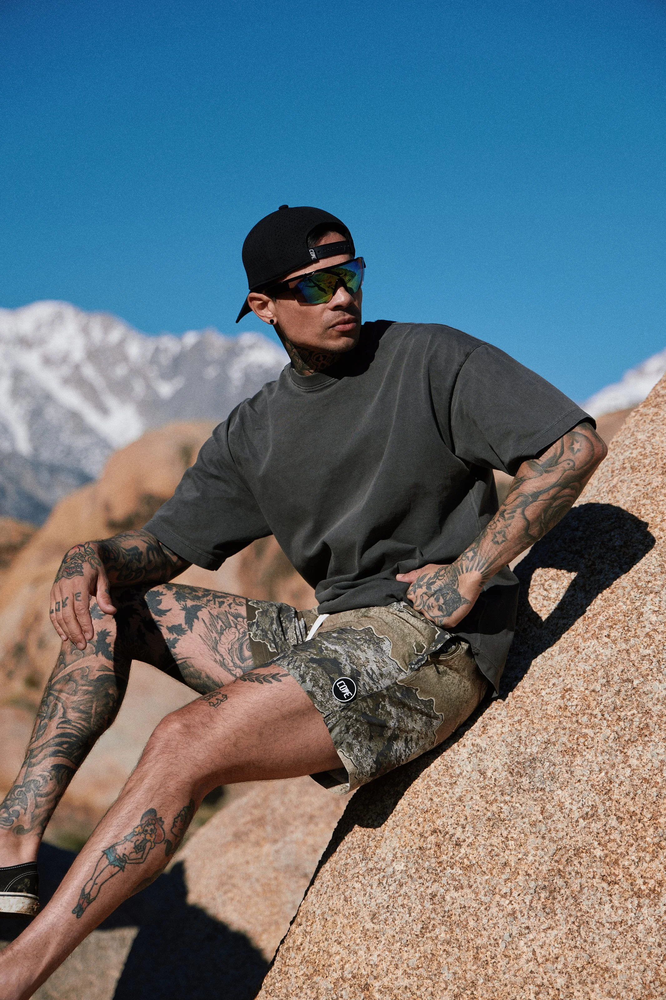 Activewear photography in Los Angeles: camo shorts, seated on boulders with snowy peaks behind, by Moises Martinez
