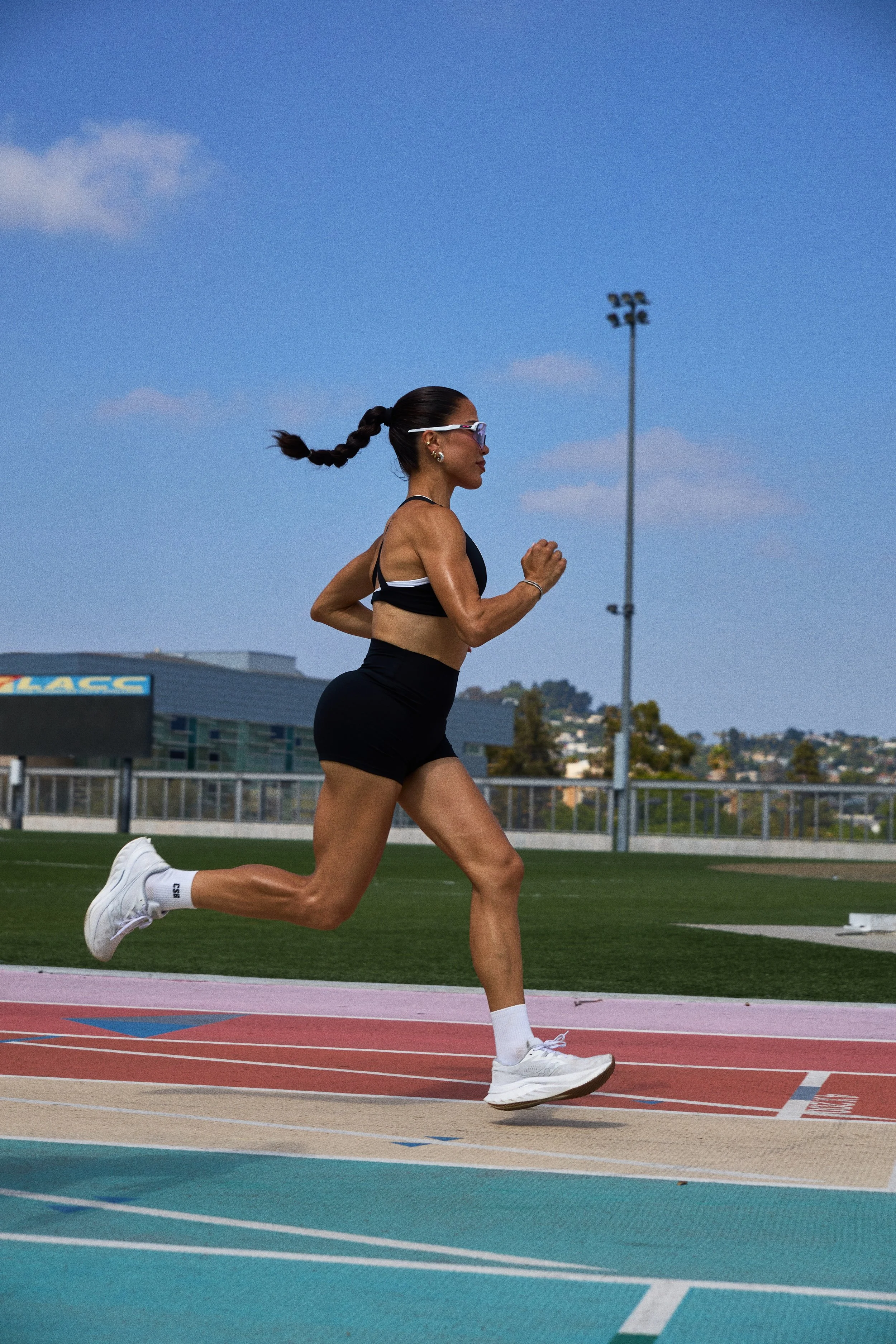 A woman running on a track at an outdoor stadium, wearing athletic clothing, sunglasses, and white running shoes.