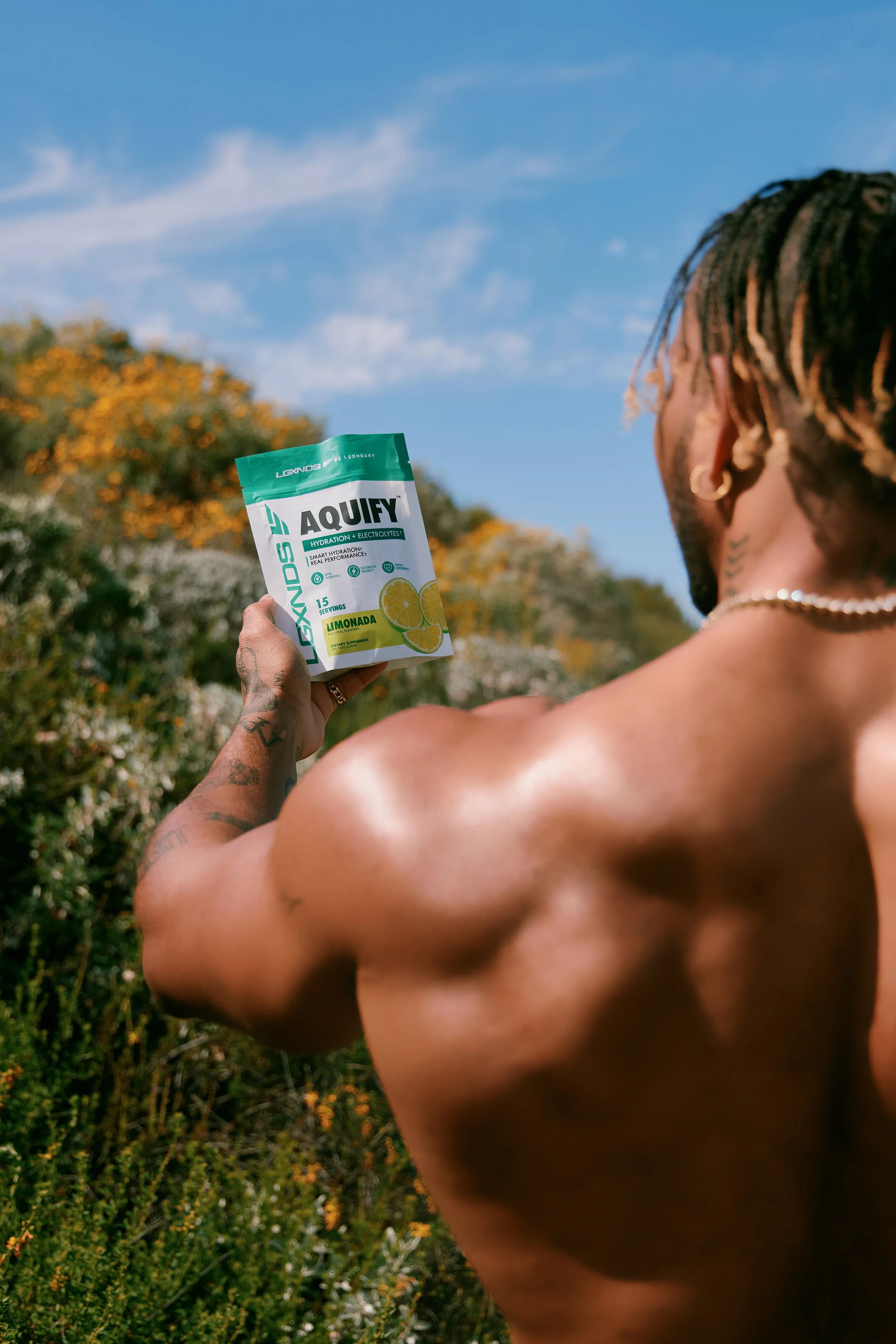 A shirtless man with tattoos and earrings holding a packet of Aqufy electrolyte powder outdoors, with a blue sky and green foliage in the background.